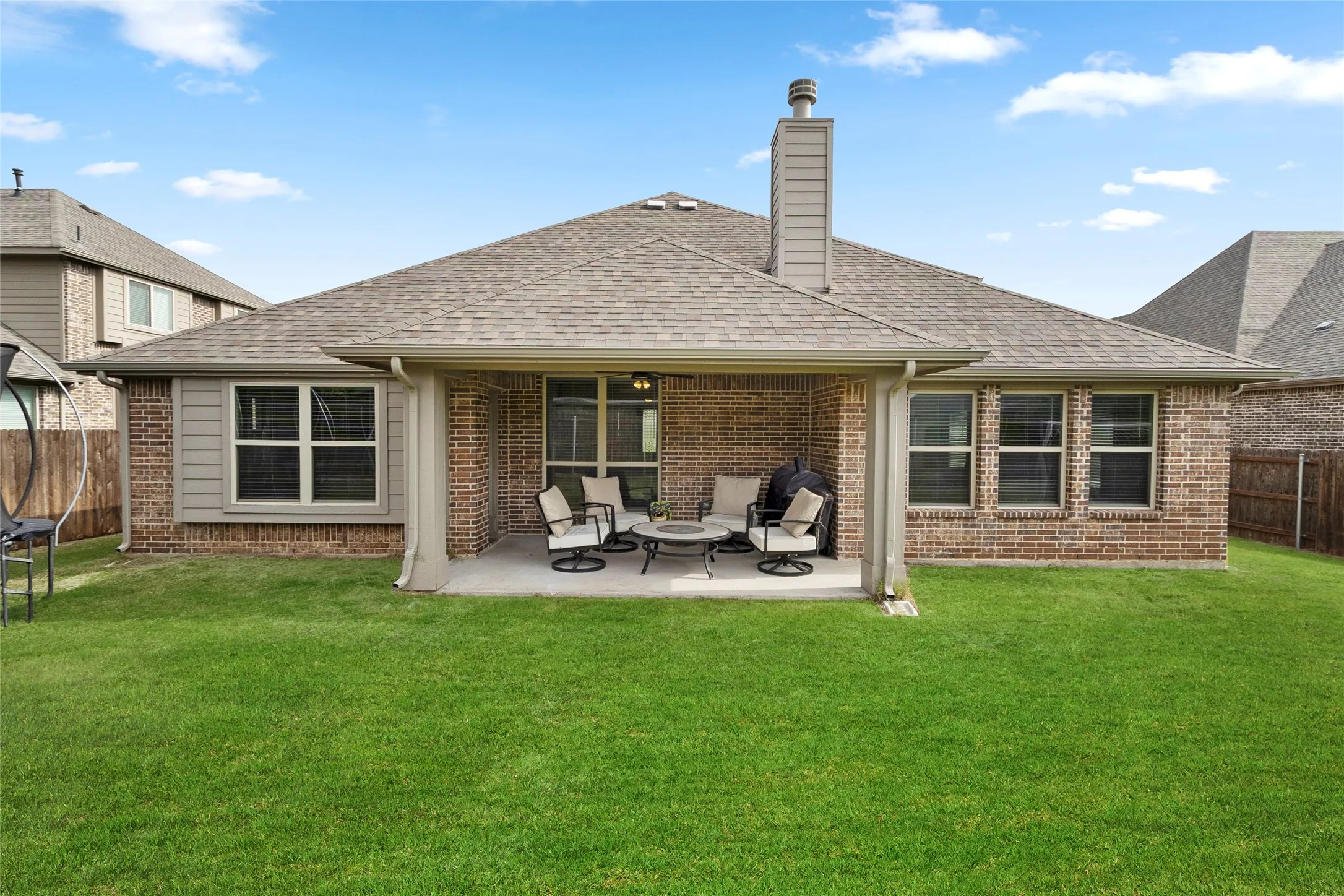 Expansive backyard featuring a covered patio, brick exterior, multiple window placements, and a pitched shingle roof