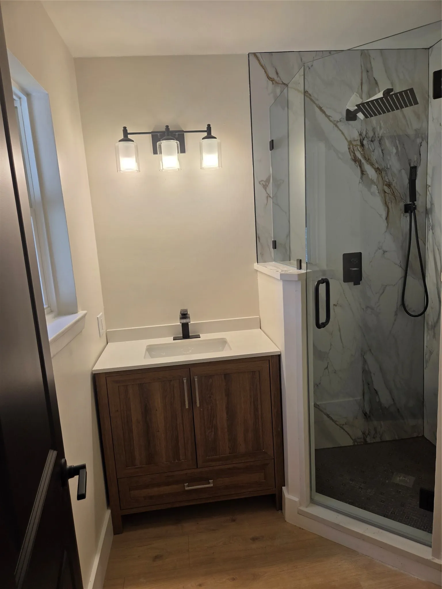 Wood-finish flooring, a contemporary vanity with an integrated sink and white countertop, and a glass-enclosed shower featuring a dark-tiled floor and a wall-mounted rain shower head