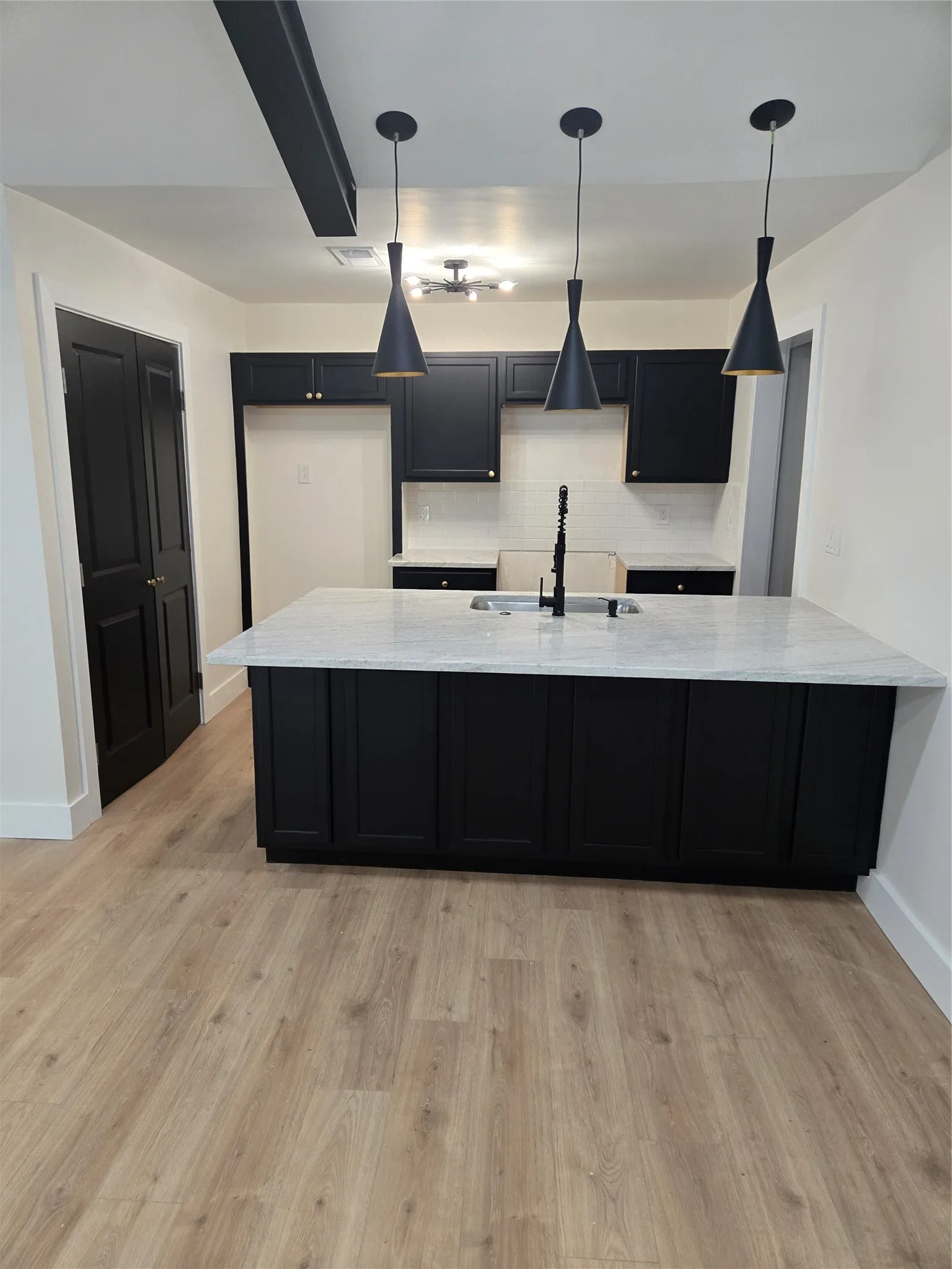 Kitchen featuring black cabinetry, a white countertop island with an integrated sink, three pendant lights, and wood-finish flooring