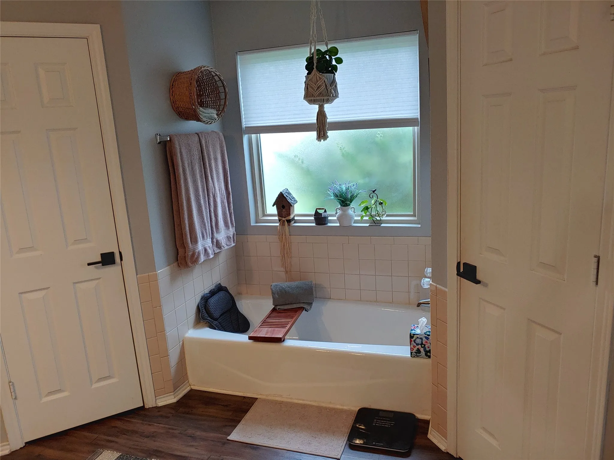 Bathroom featuring a white bathtub, tiled surround, window with blind, wood-finish flooring, and two paneled doors with black hardware