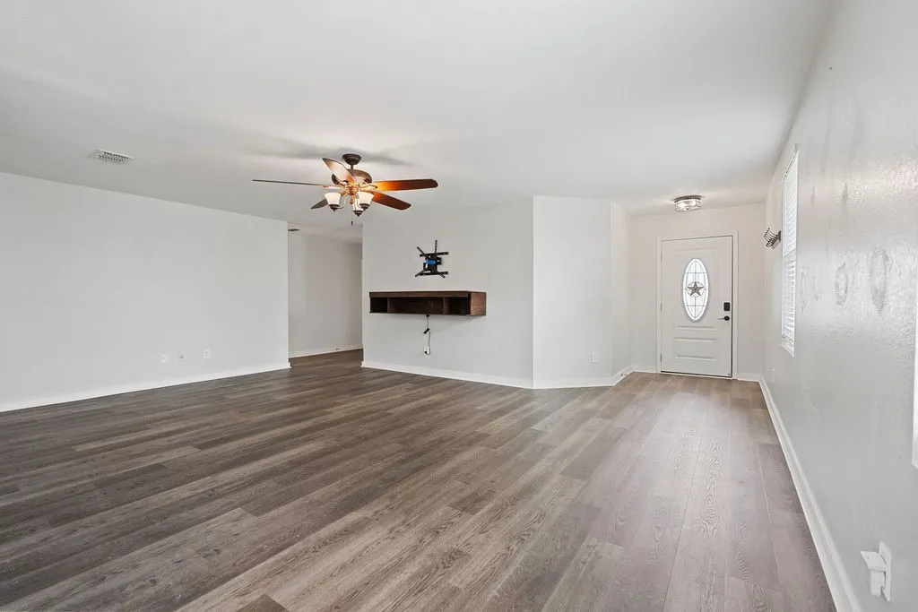 Spacious living area featuring wood-finish flooring, a ceiling fan with integrated lighting, a built-in floating shelf, a front door with an oval glass insert, and a window with blinds