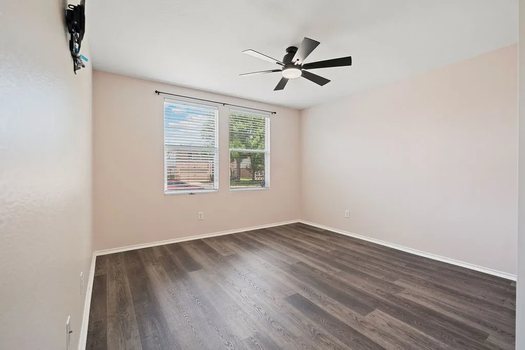 Spacious room featuring wood-finish flooring, two windows with blinds, a ceiling fan with integrated lighting, and neutral wall tones