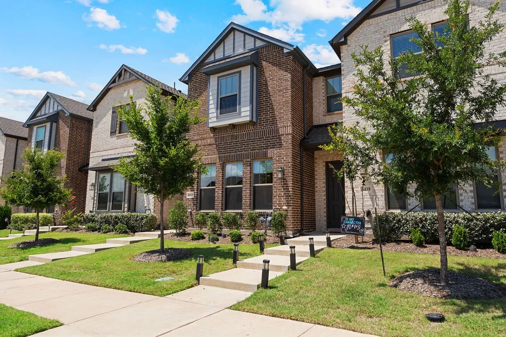Contemporary brick and stone facade featuring gabled roofs, multiple windows, established landscaping, a concrete walkway, and decorative path lighting