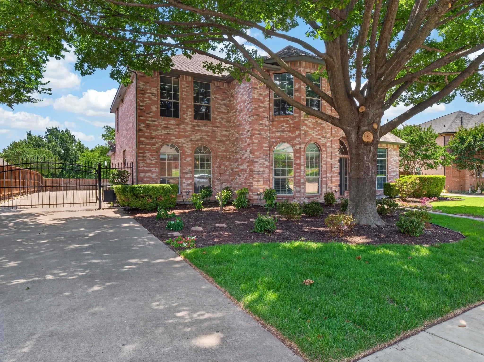 Brick facade residence with a prominent mature tree, featuring arched windows on the lower level, a manicured lawn, and a private driveway with an ornate metal gate