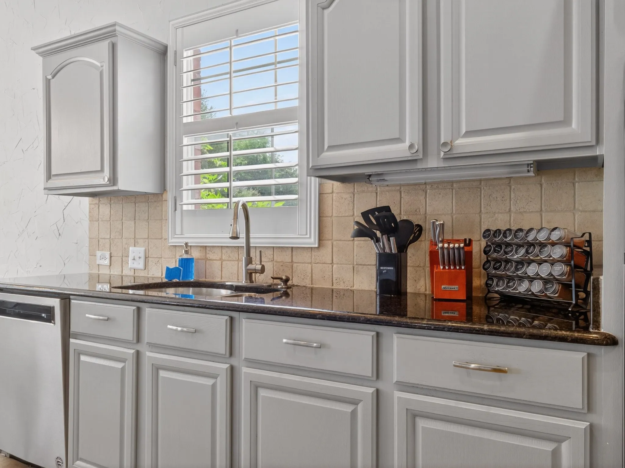 Kitchen featuring light gray cabinetry with brushed nickel hardware, dark countertop, square tile backsplash, and a stainless steel dishwasher