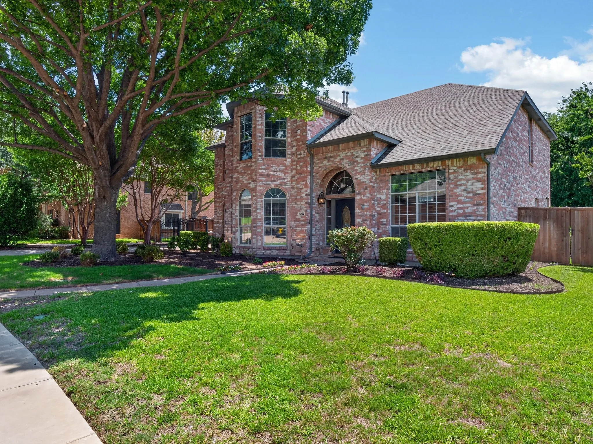 Brick exterior featuring an arched entryway, multiple large windows, a mature shade tree, a well-maintained lawn, and established shrubbery