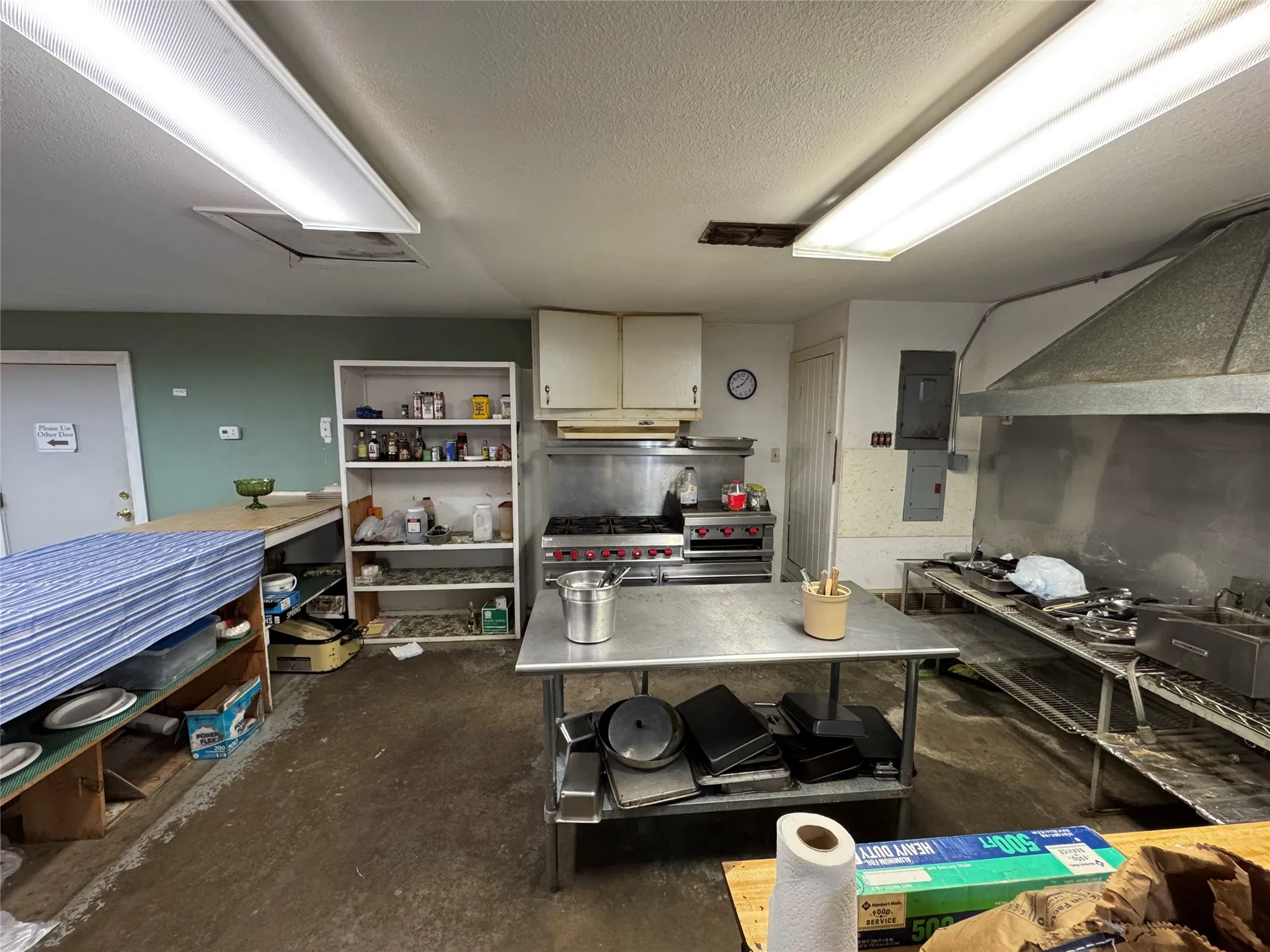 Commercial kitchen space featuring a stainless steel range with overhead ventilation, multiple stainless steel work tables, and extensive shelving