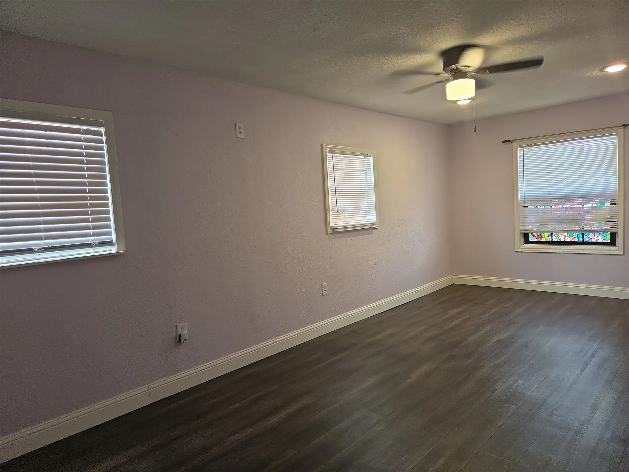 Spacious room featuring wood-finish flooring, light purple wall paint, white baseboards, and multiple windows with blinds