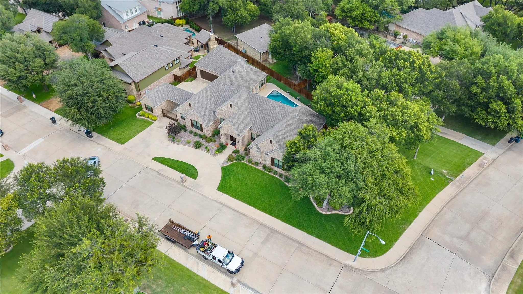 Expansive corner lot featuring a brick and stone exterior, a concrete driveway with an attached garage, a private swimming pool, and mature trees providing natural screening