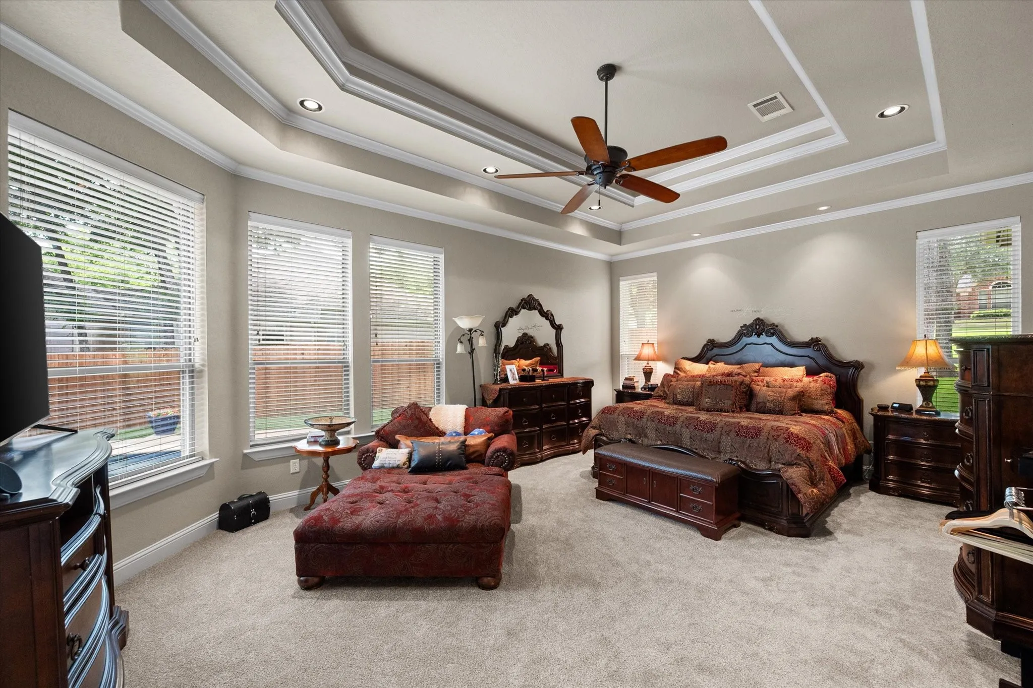 Spacious bedroom featuring a tray ceiling with recessed lighting and a ceiling fan, multiple windows with blinds, neutral wall paint, and plush carpet flooring