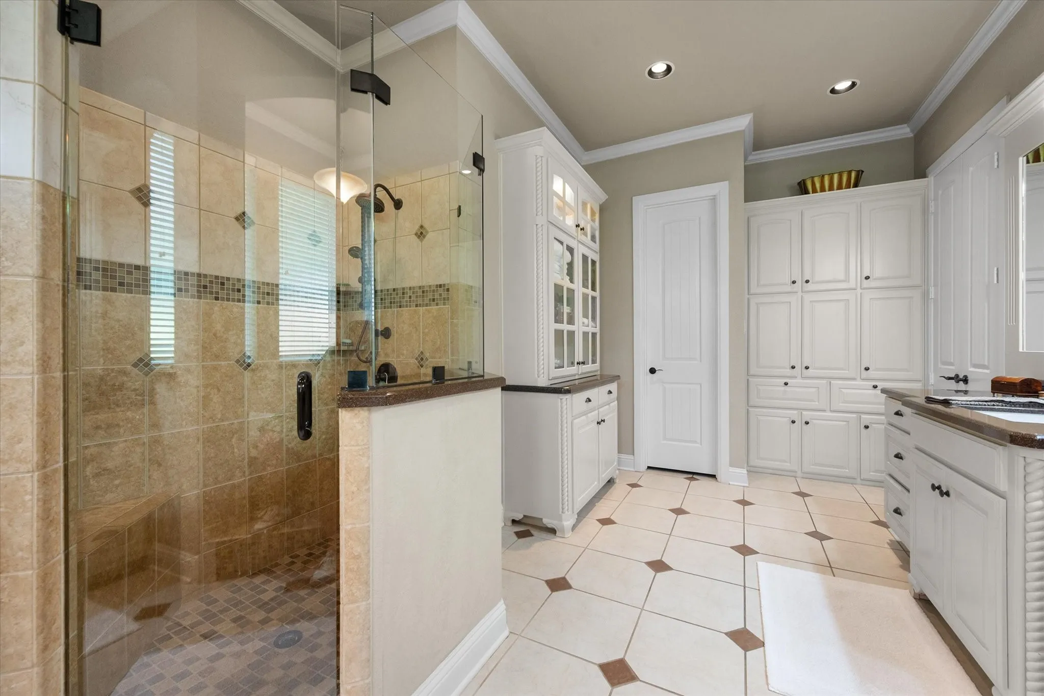 Spacious bathroom featuring a frameless glass shower enclosure with floor-to-ceiling tile, extensive built-in cabinetry, recessed lighting, and tile flooring with accent inserts