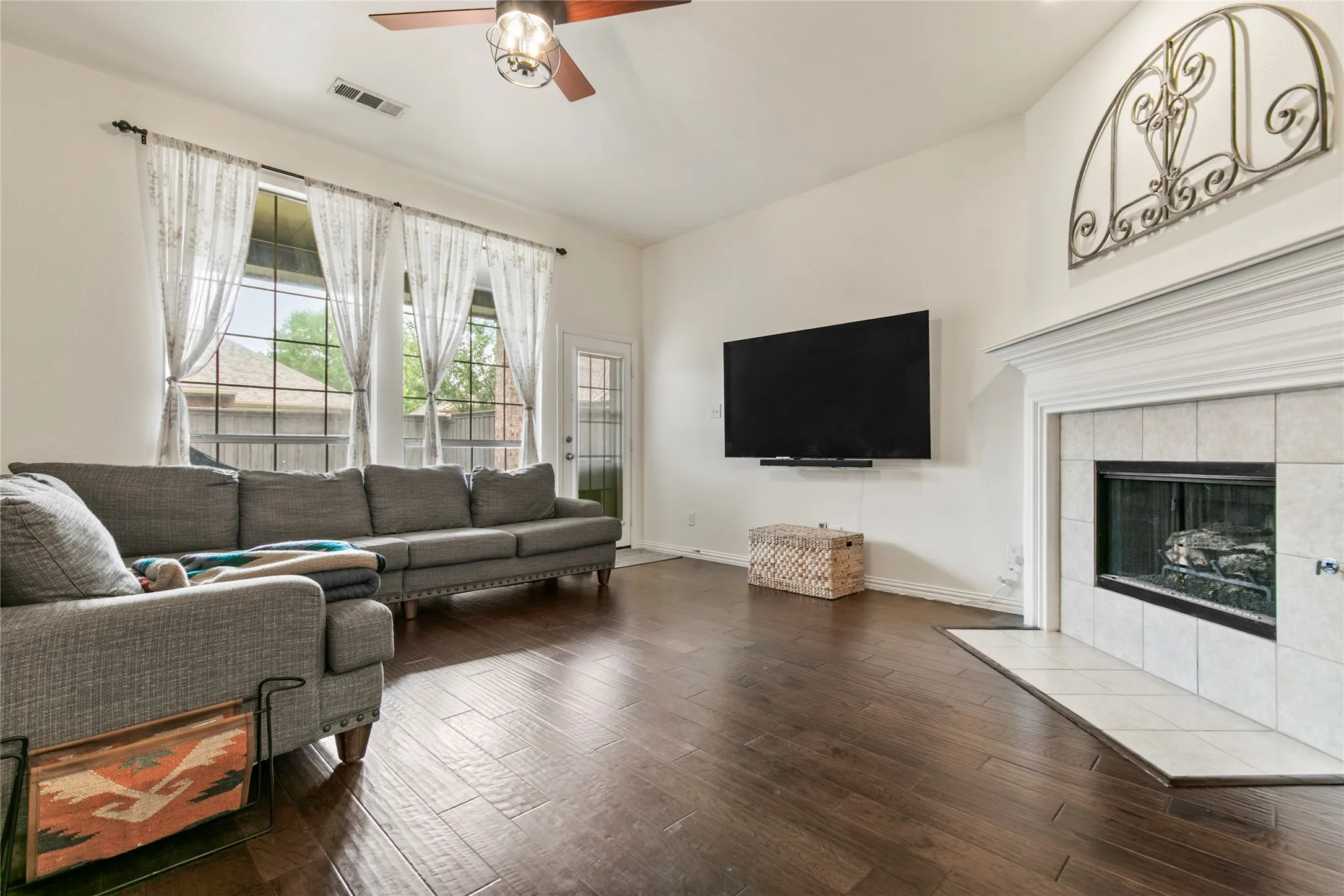 Spacious living area featuring wood-finish flooring, a tiled fireplace with a classic mantel, and a ceiling fan with integrated lighting