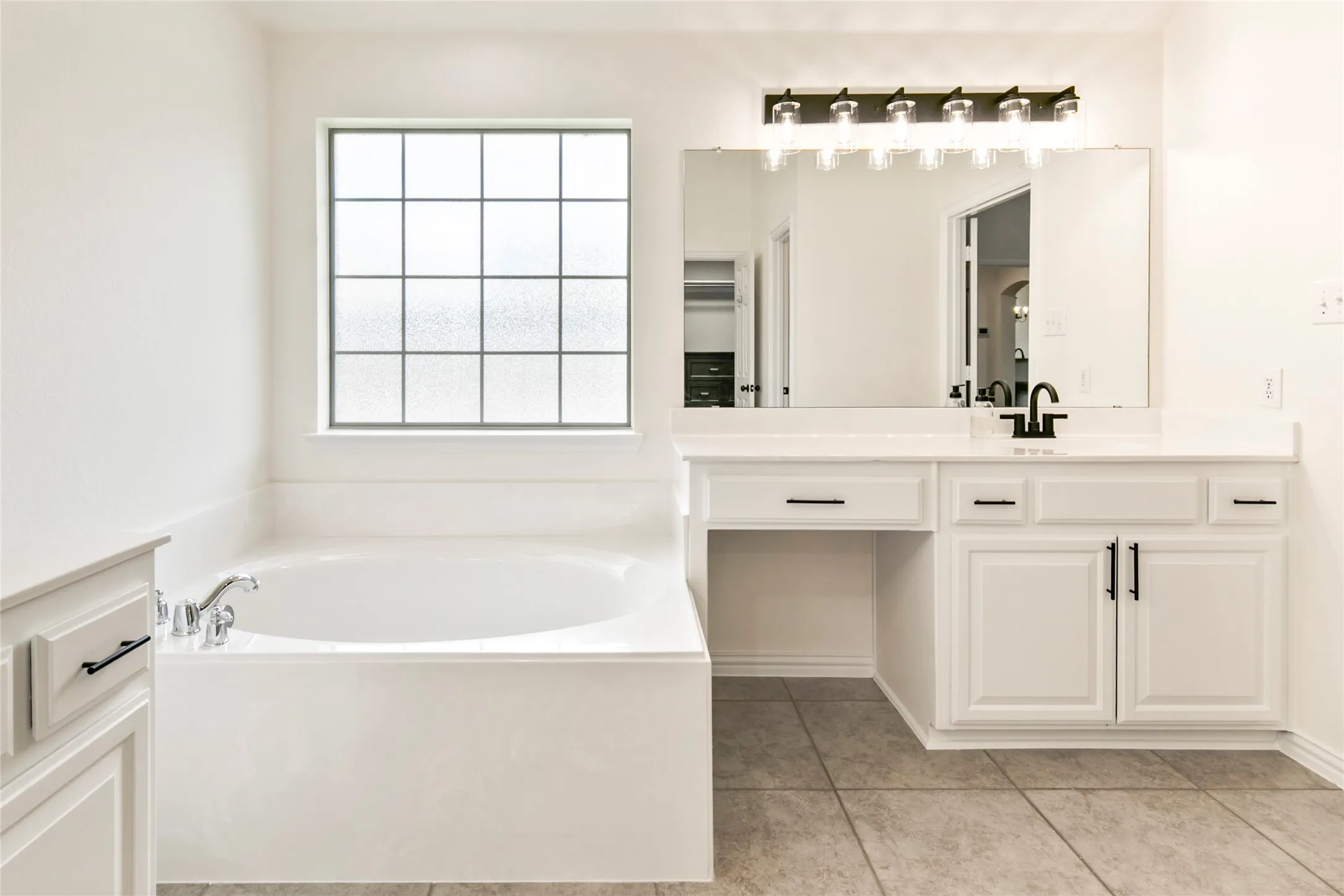 White soaking tub with chrome fixtures, frosted window, and a vanity with a white countertop, black faucet, and sconce lighting