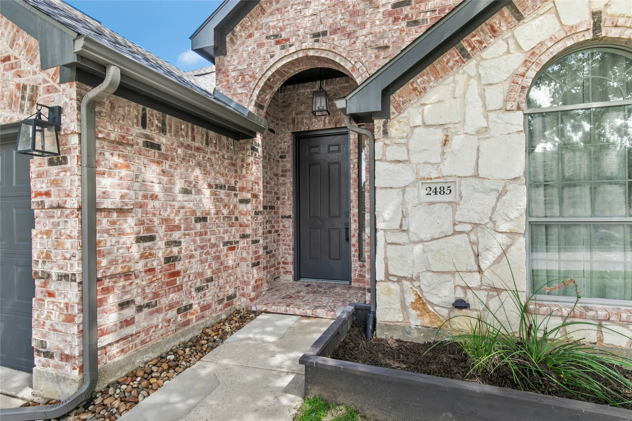 Exterior facade featuring a combination of brick and natural stone, arched entryway, dark-finish front door, arched window, and integrated gutter system