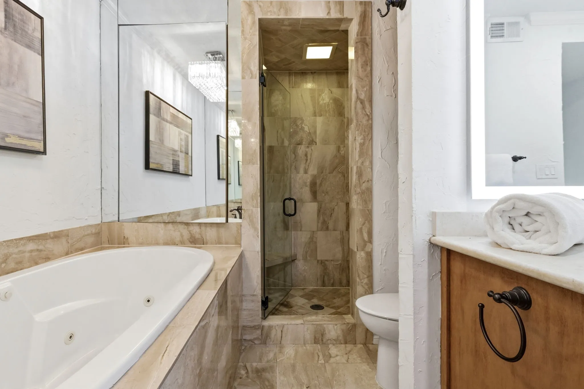 Bathroom featuring a built-in jetted tub, a glass-enclosed shower, natural stone tile, an illuminated mirror, and a wood-finish vanity with a stone countertop
