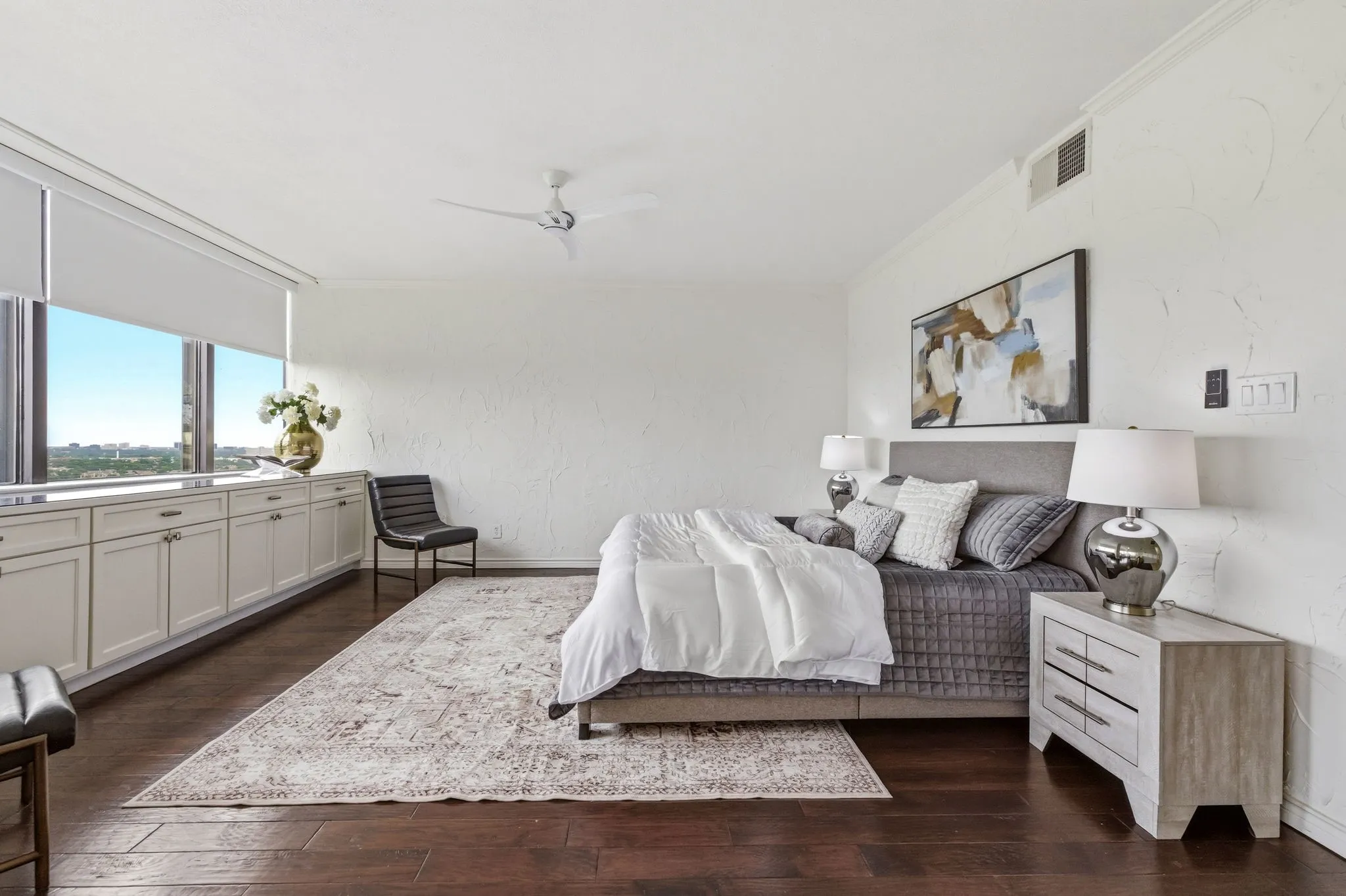 Spacious bedroom featuring dark wood-finish flooring, a full wall of floor-to-ceiling windows with roller shades, and a lengthy built-in cabinet unit with a light-toned countertop