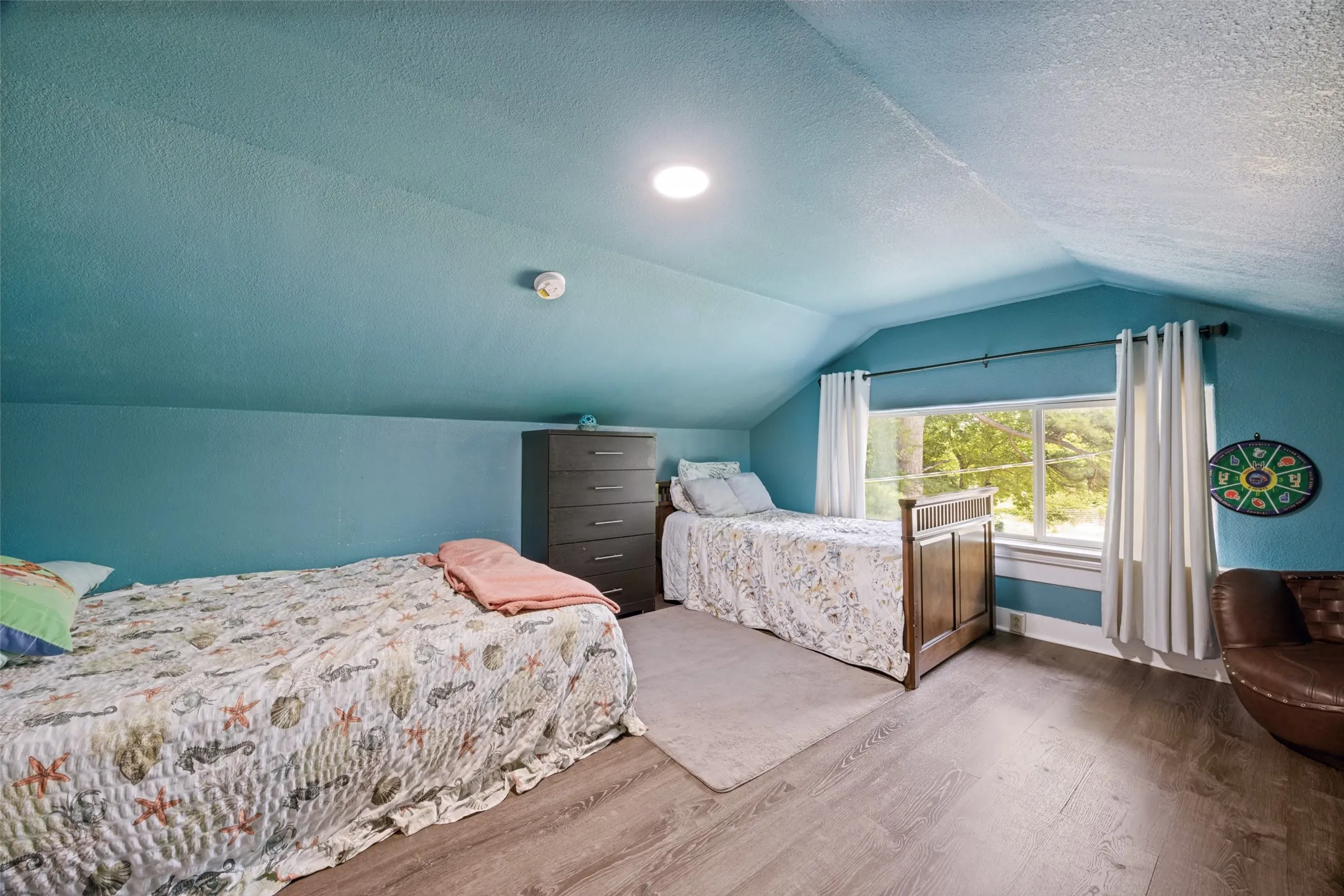 Sloped ceiling room featuring wood-finish flooring, recessed lighting, and a prominent window with white curtains