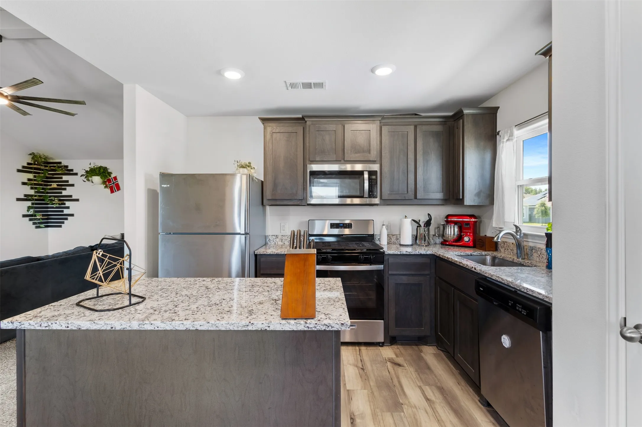 Modern kitchen featuring a central island, light-toned wood-finish flooring, stainless steel appliances, dark wood cabinetry, and granite countertops