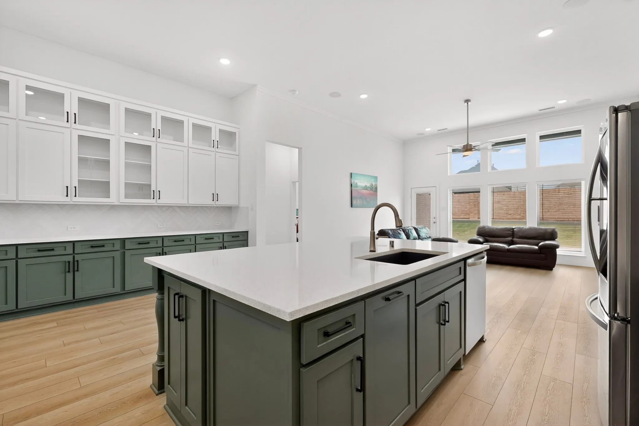 Kitchen featuring a large central island with a built-in sink and white countertop, complemented by dark green base cabinetry and white upper cabinetry with glass inserts