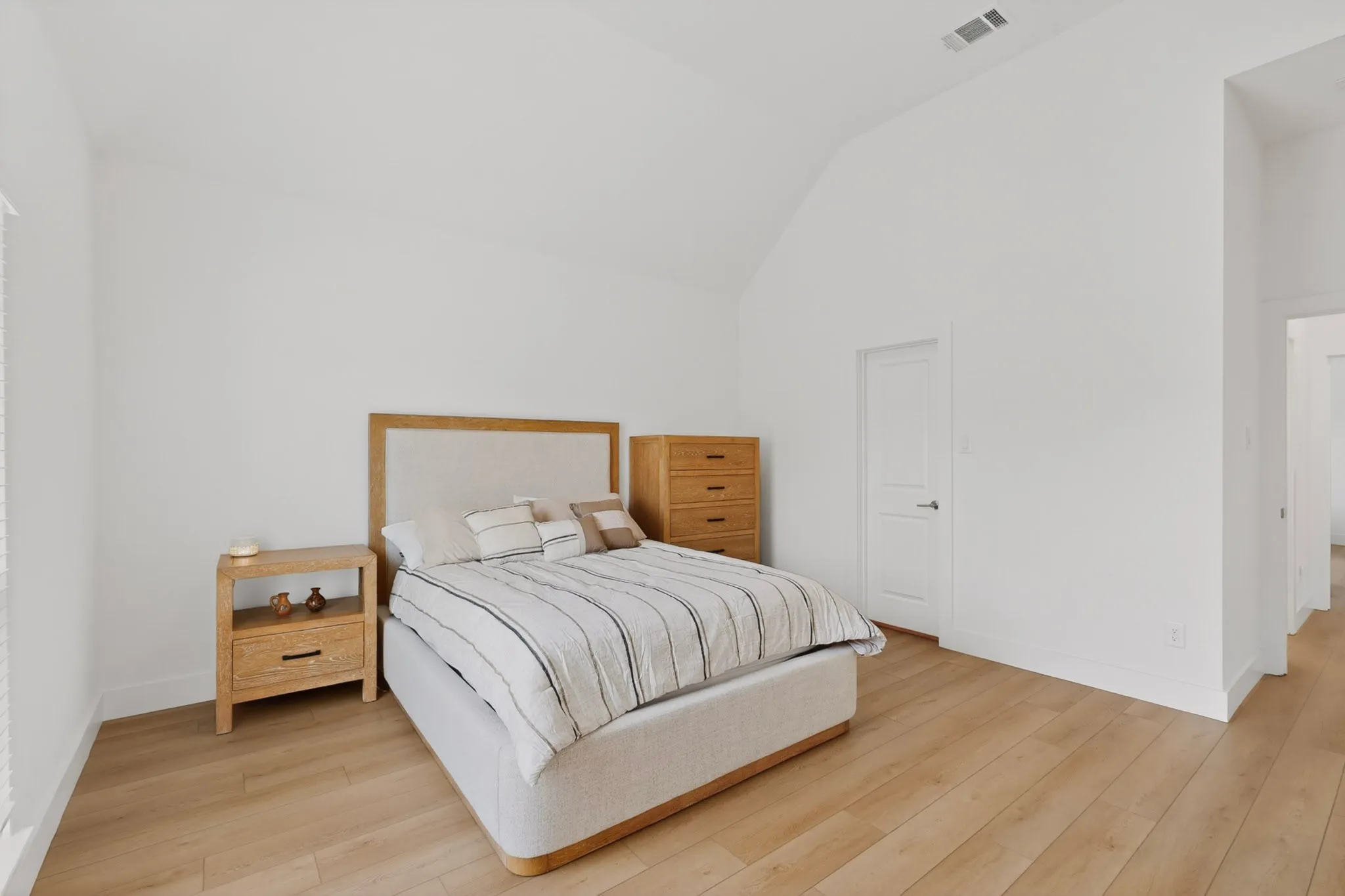 Bright bedroom featuring vaulted ceilings, light wood-finish flooring, white baseboards, and a white door with contemporary hardware