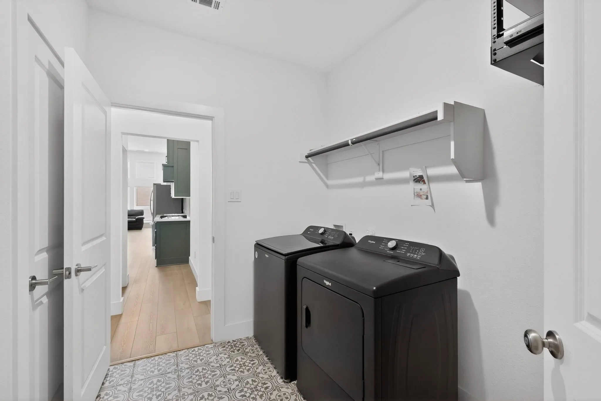 Dedicated laundry area featuring patterned floor tile, overhead shelving, and light wood-finish flooring visible beyond the doorway
