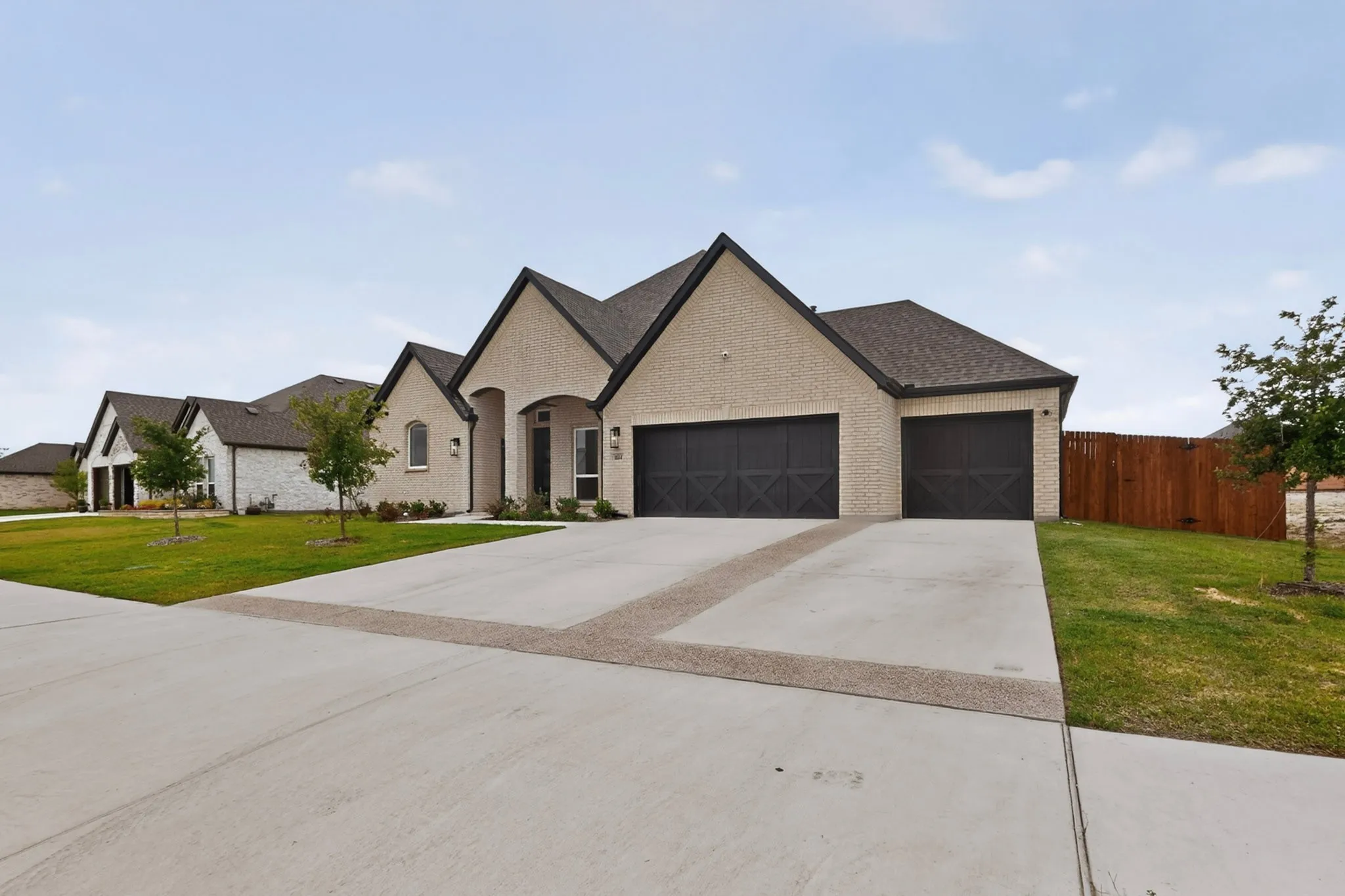 Cream brick exterior featuring dark trim, a two-car garage with X-brace doors, a covered entryway, and a concrete driveway with decorative banding