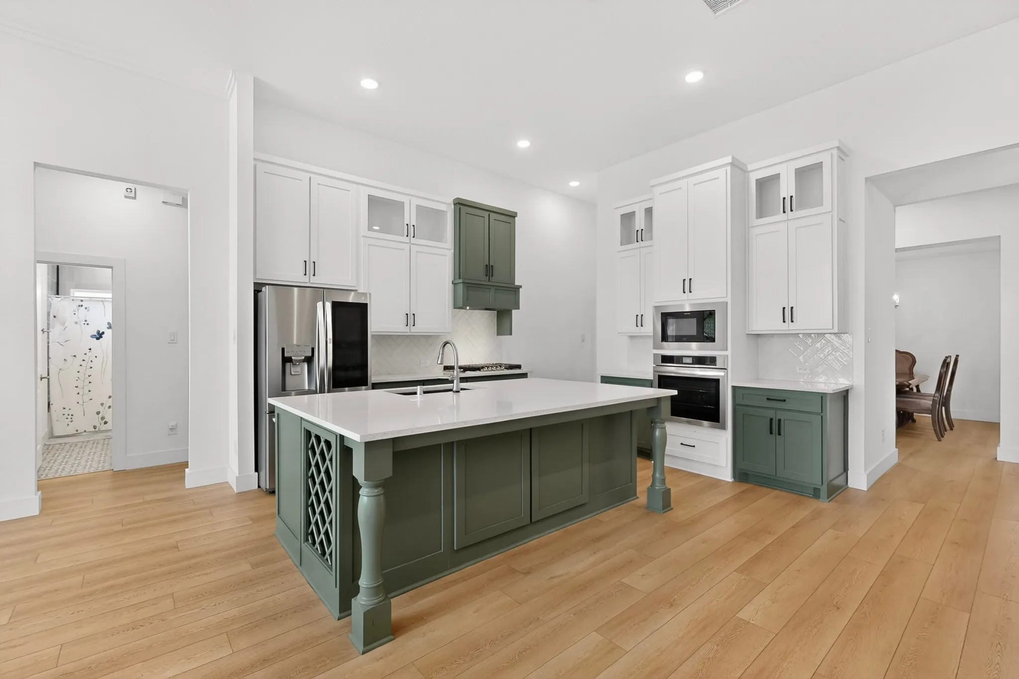 Bright kitchen featuring a central island with white countertop, stainless steel appliances, white cabinetry with glass inserts, green lower cabinetry, and wood-finish flooring