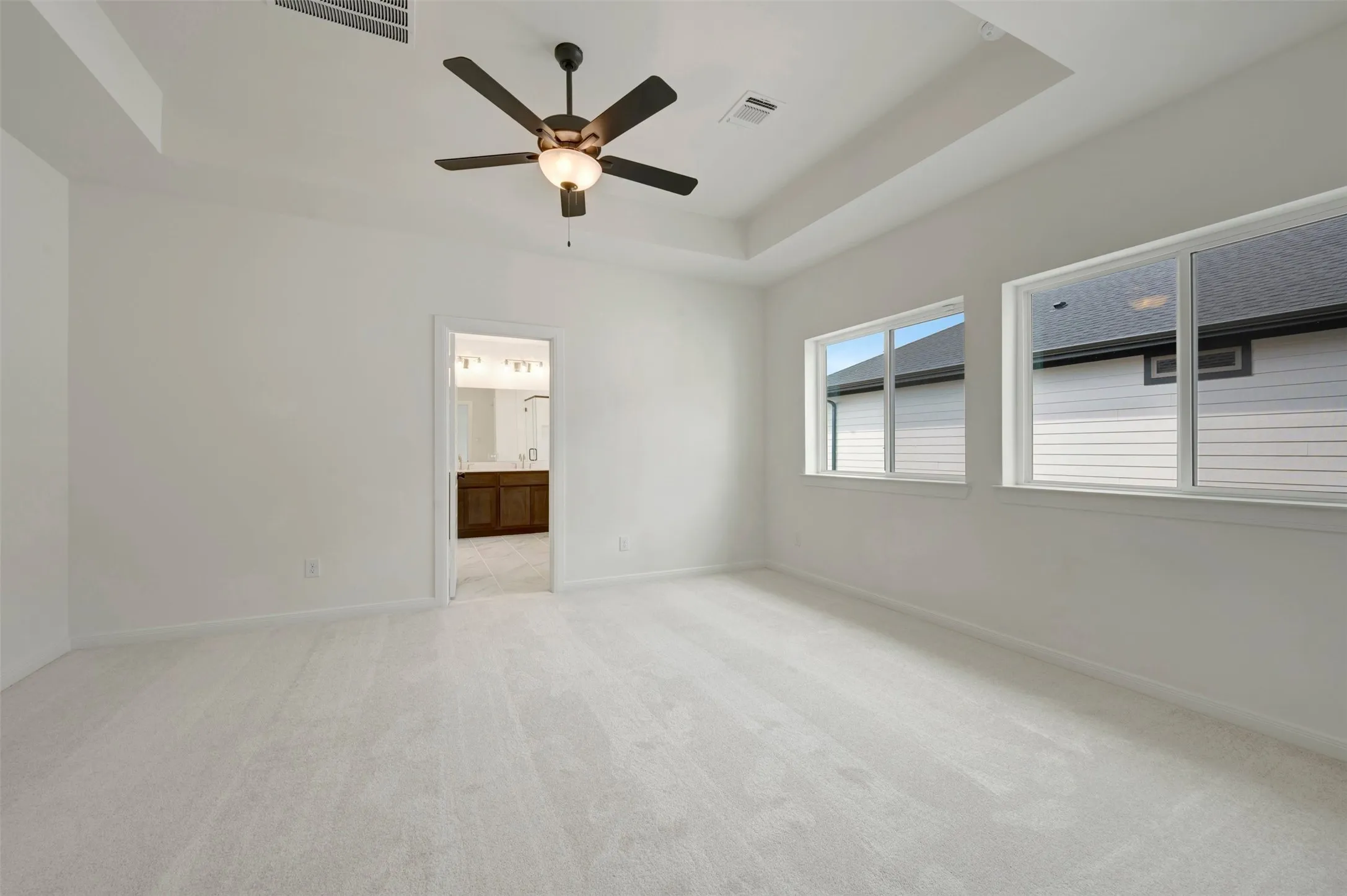 Tray ceiling, ceiling fan with integrated lighting, two windows, wall-to-wall carpeting, and an adjoining bathroom with a wood-finish vanity