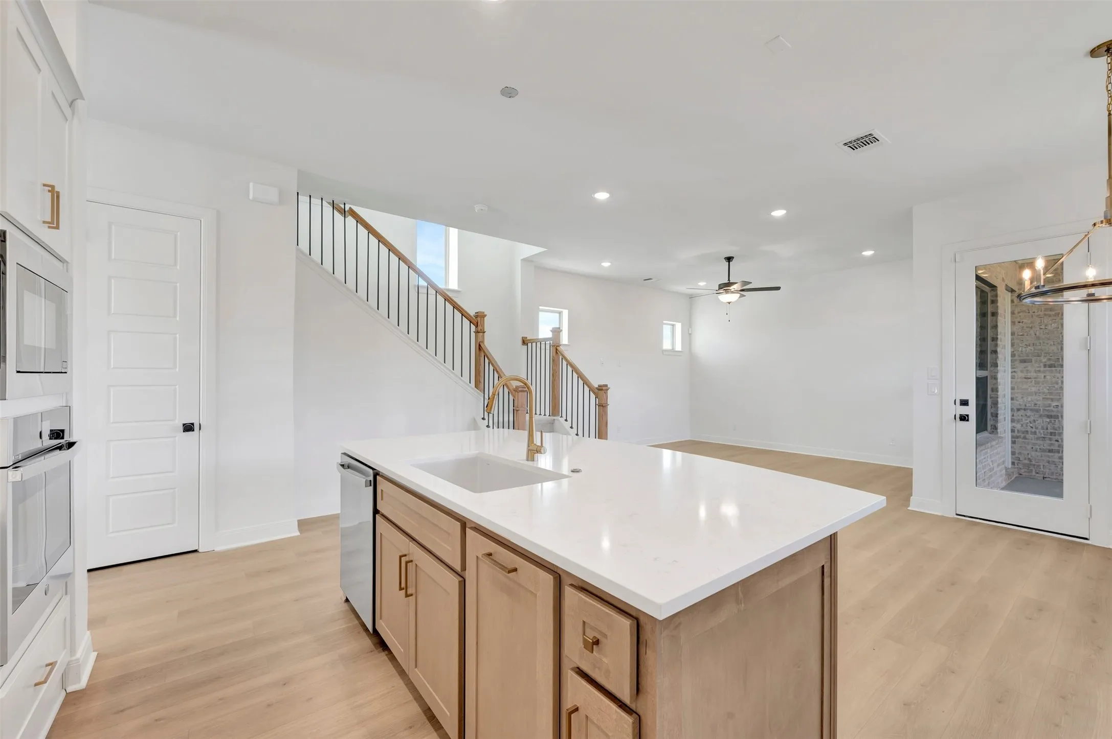 Kitchen island with a white countertop and undermount sink, built-in stainless steel dishwasher, wood-finish flooring, staircase with dark metal balusters, and white paneled doors