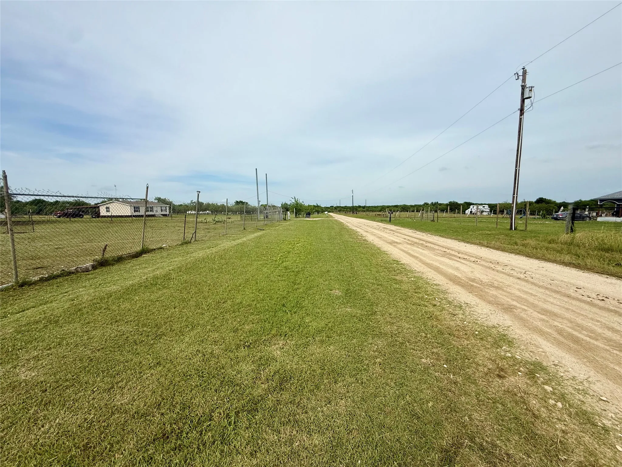 Expansive grassy lot with a dirt road, featuring a chain-link fence and utility poles