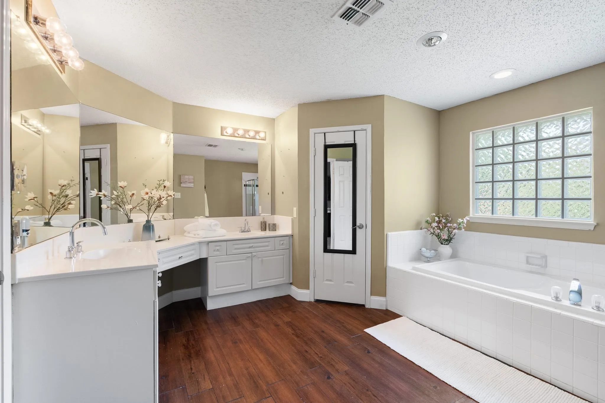 Bathroom featuring wood-finish flooring, a built-in soaking tub with white tile surround, and a glass block window