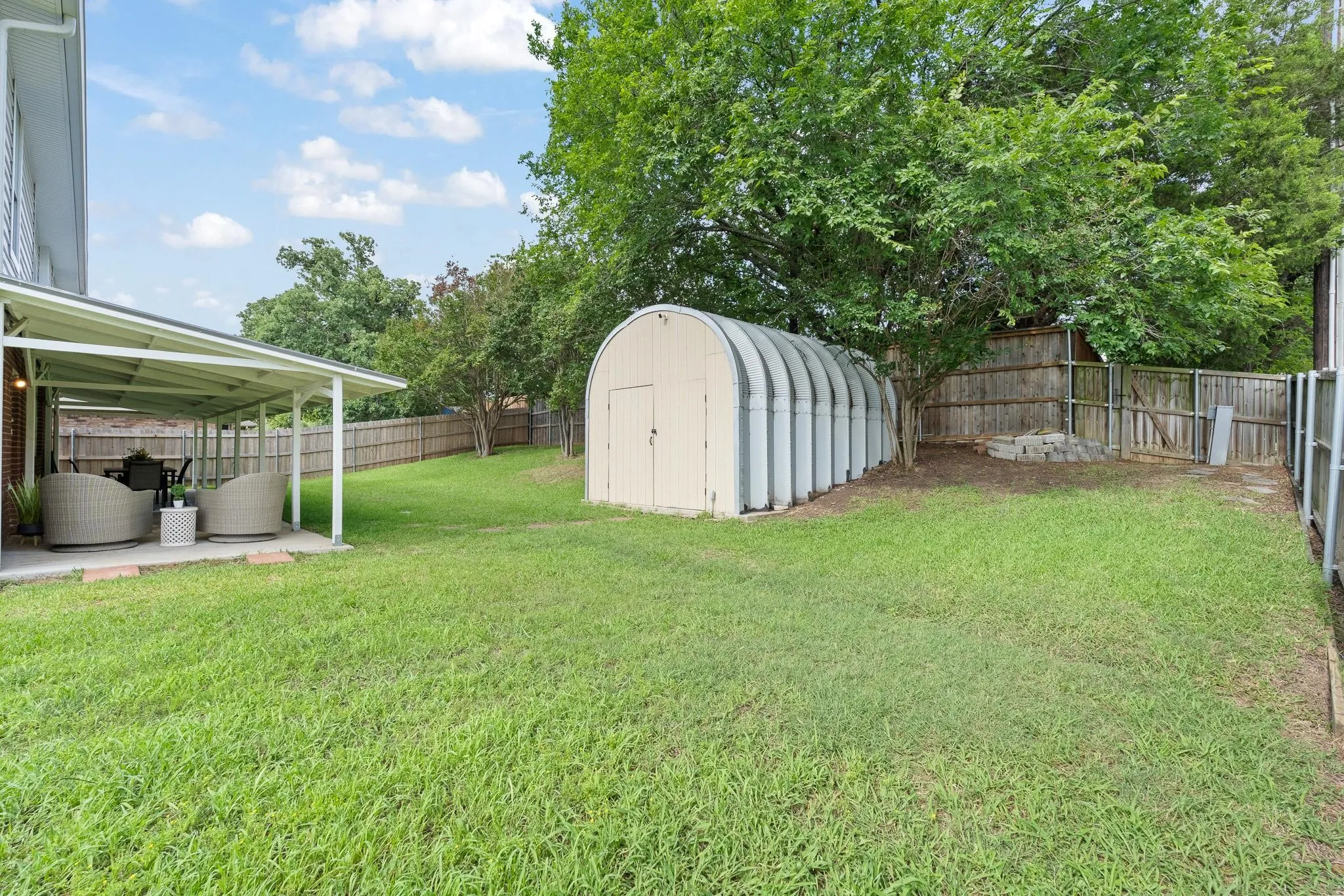 Expansive grass yard featuring a covered patio, a large storage shed, mature trees, and privacy fencing