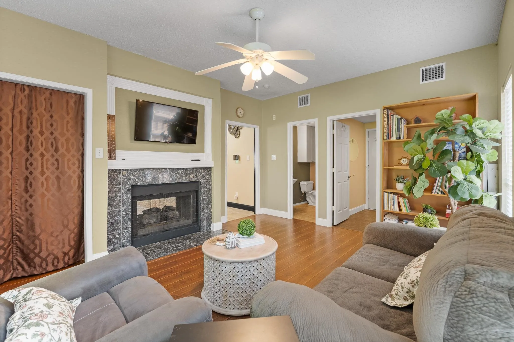 Family room featuring wood-finish flooring, a ceiling fan, and a fireplace with a stone surround