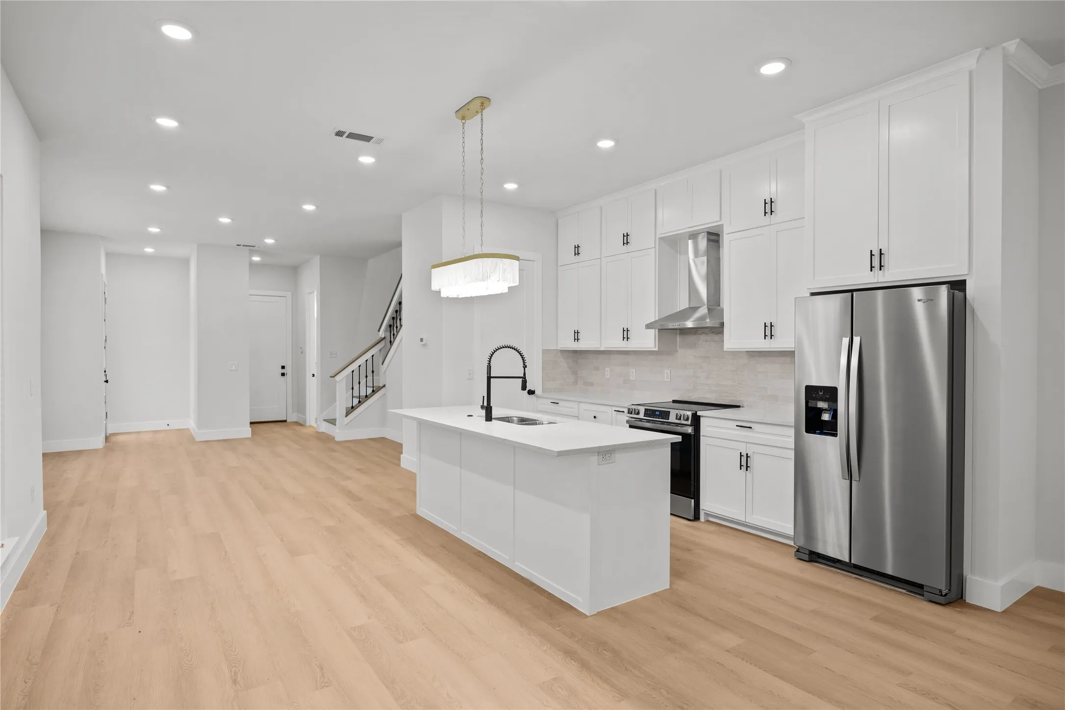 Contemporary kitchen featuring an island with a built-in sink, white cabinetry with dark hardware, stainless steel appliances, light-toned wood-finish flooring, and recessed lighting