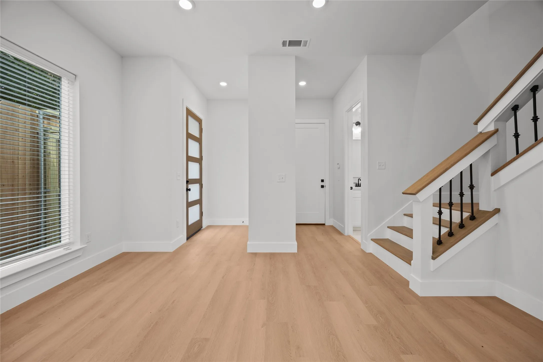 Open-concept living area featuring light wood-finish flooring, recessed lighting, a modern entry door with frosted glass panels, a staircase with wood handrails and black metal balusters, and a powder room with a vanity and wall-mounted light fixture