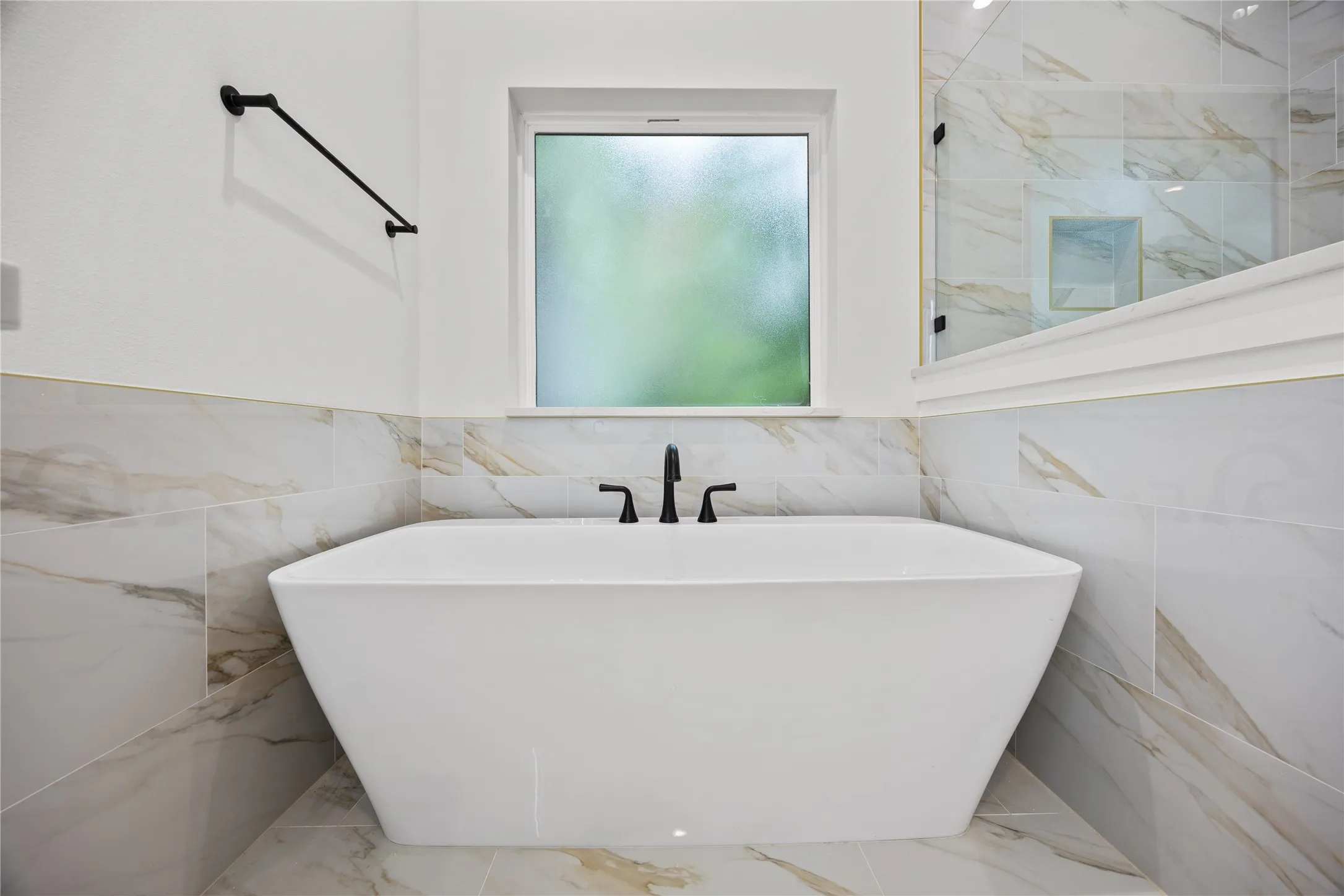 Freestanding soaking tub with matte black fixtures, set against a backdrop of large format marble-look tile walls and flooring