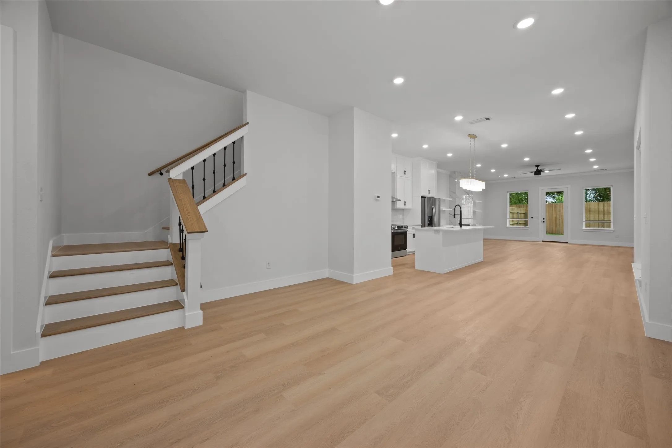 Open-concept interior featuring light wood-finish flooring throughout, a modern staircase with wood handrails and black balusters, recessed lighting, a kitchen island with a dark faucet, and white kitchen cabinetry