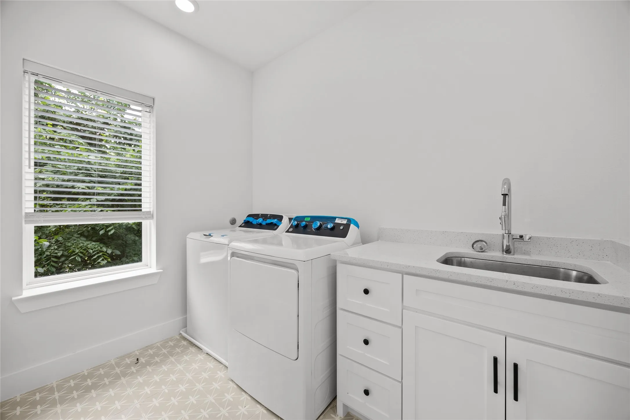 Utility room featuring a window with blinds, a built-in sink with a gooseneck faucet, white shaker-style cabinetry, a light-toned patterned floor, and a recessed ceiling light