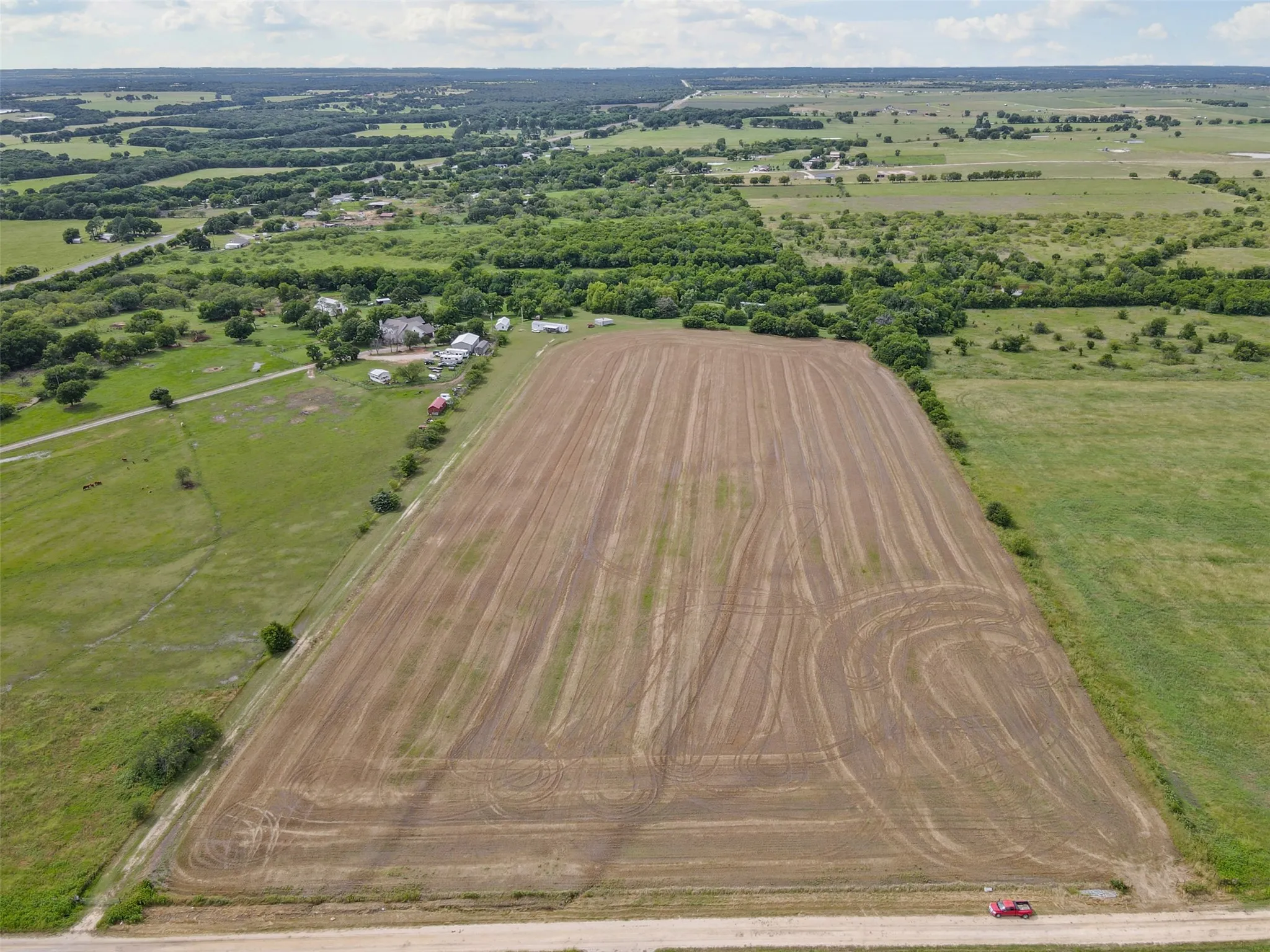 Expansive agricultural field with tilled soil, bordered by mature trees and verdant pastureland