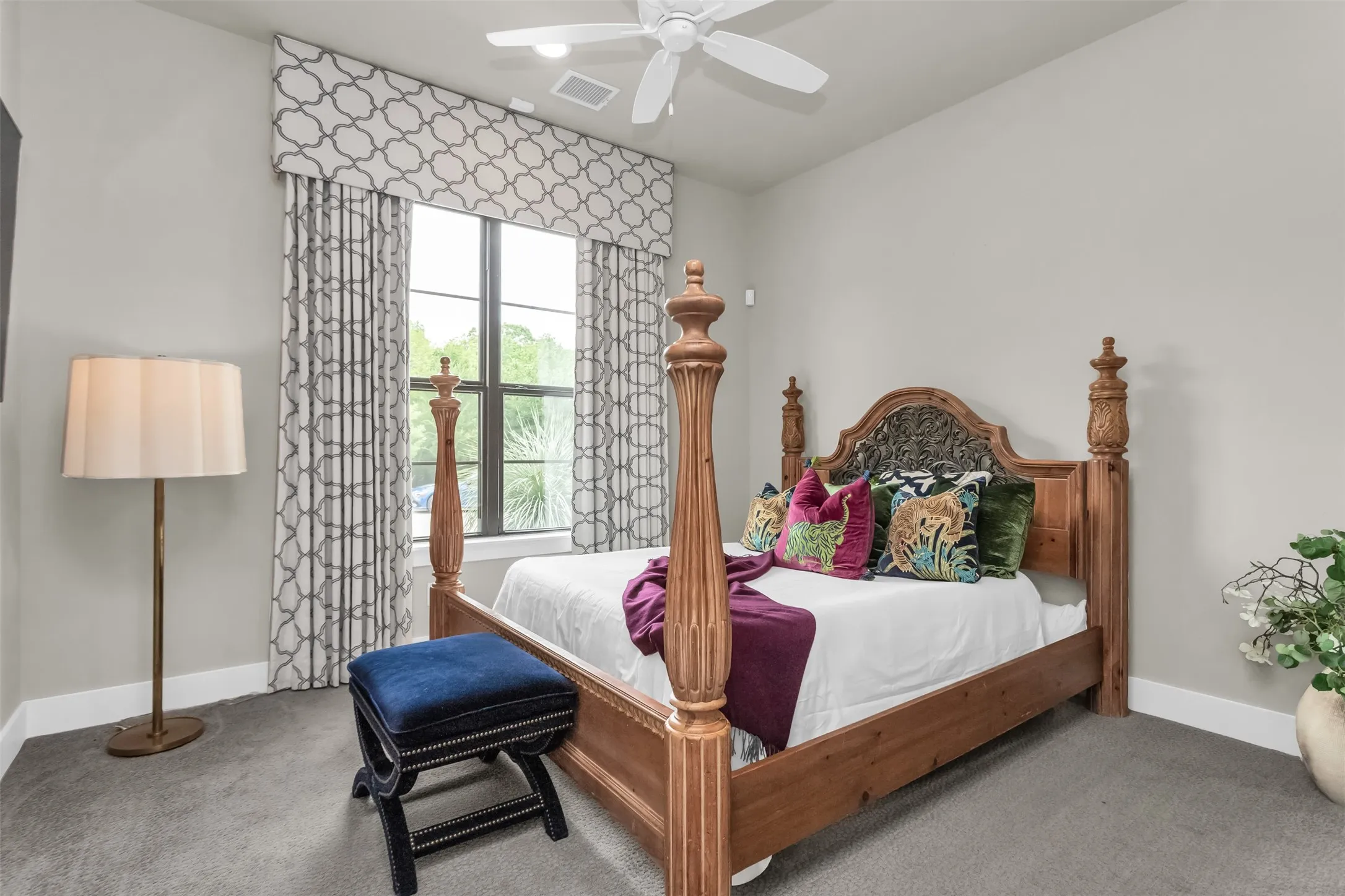 Carpeted bedroom featuring a ceiling fan with integrated lighting, a large window with patterned drapes, light gray wall paint, and white baseboards