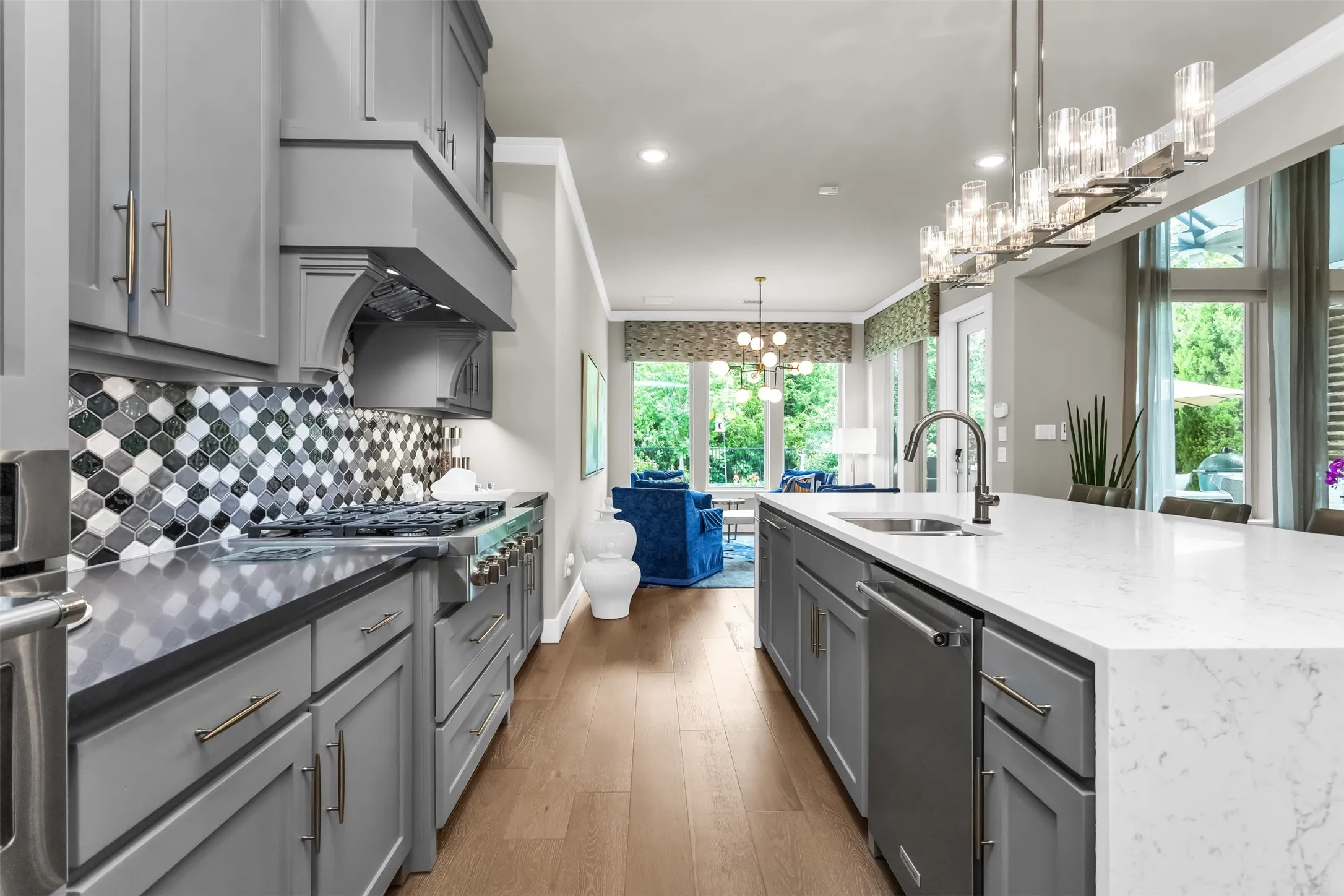 Gourmet kitchen featuring grey cabinetry, stainless steel range with custom hood, mosaic tile backsplash, spacious center island with sink, and wood-finish flooring