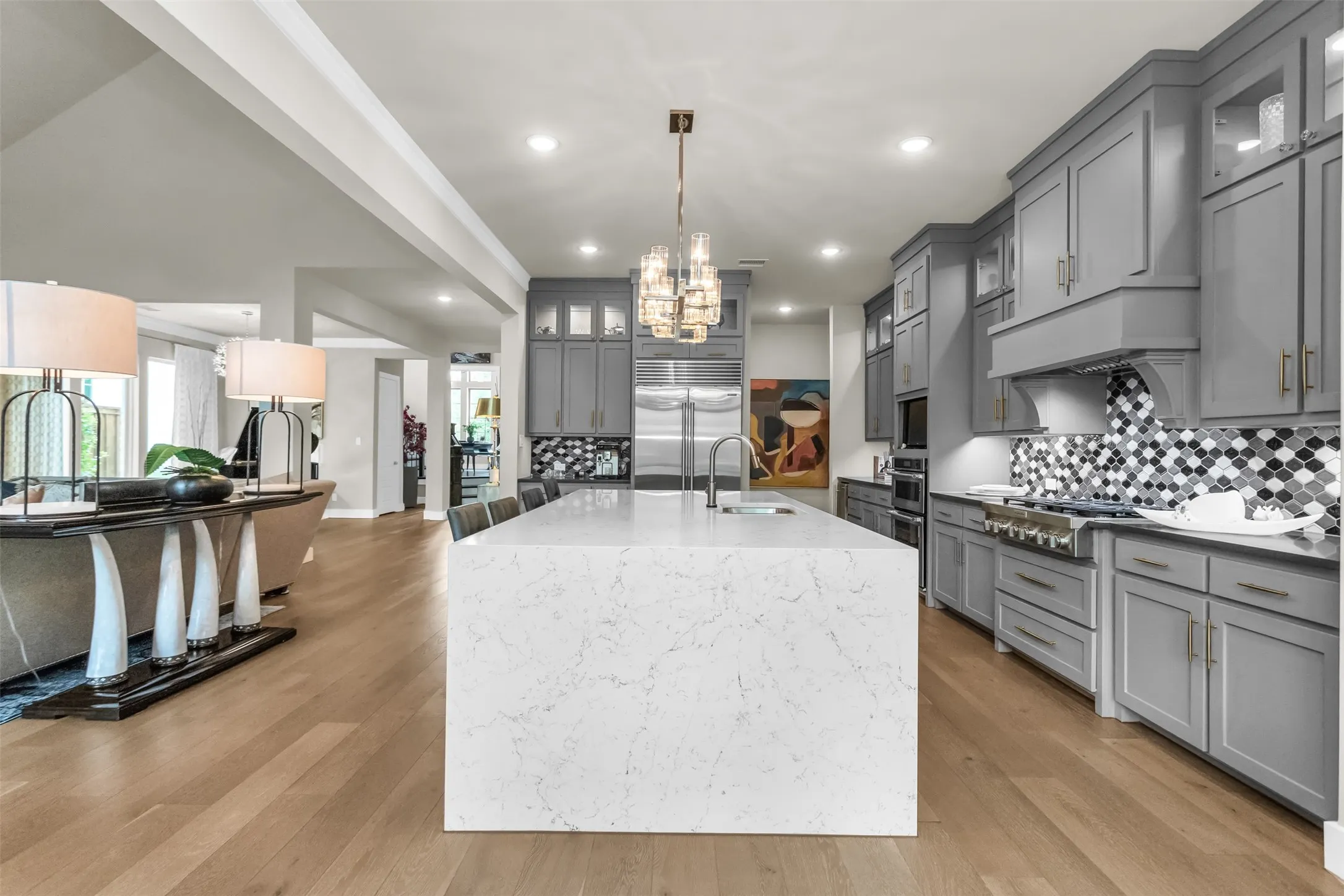 Gourmet kitchen featuring a waterfall island with a built-in sink, gray cabinetry, a decorative range hood, stainless steel appliances, and wood-finish flooring