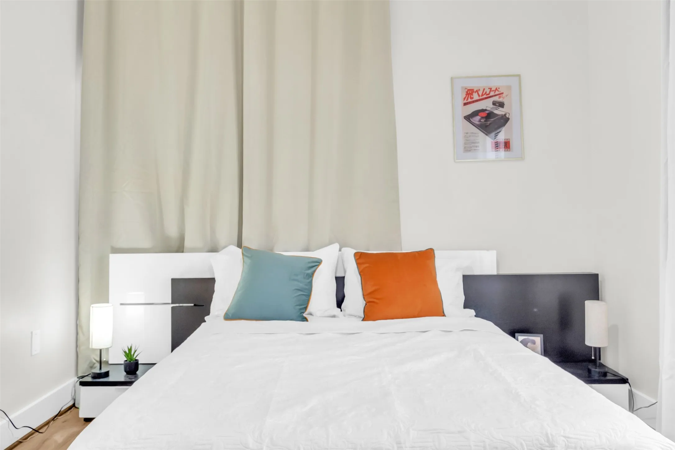 Bedroom featuring a white bed frame with integrated side tables, light-colored privacy drapes, and wood-finish flooring