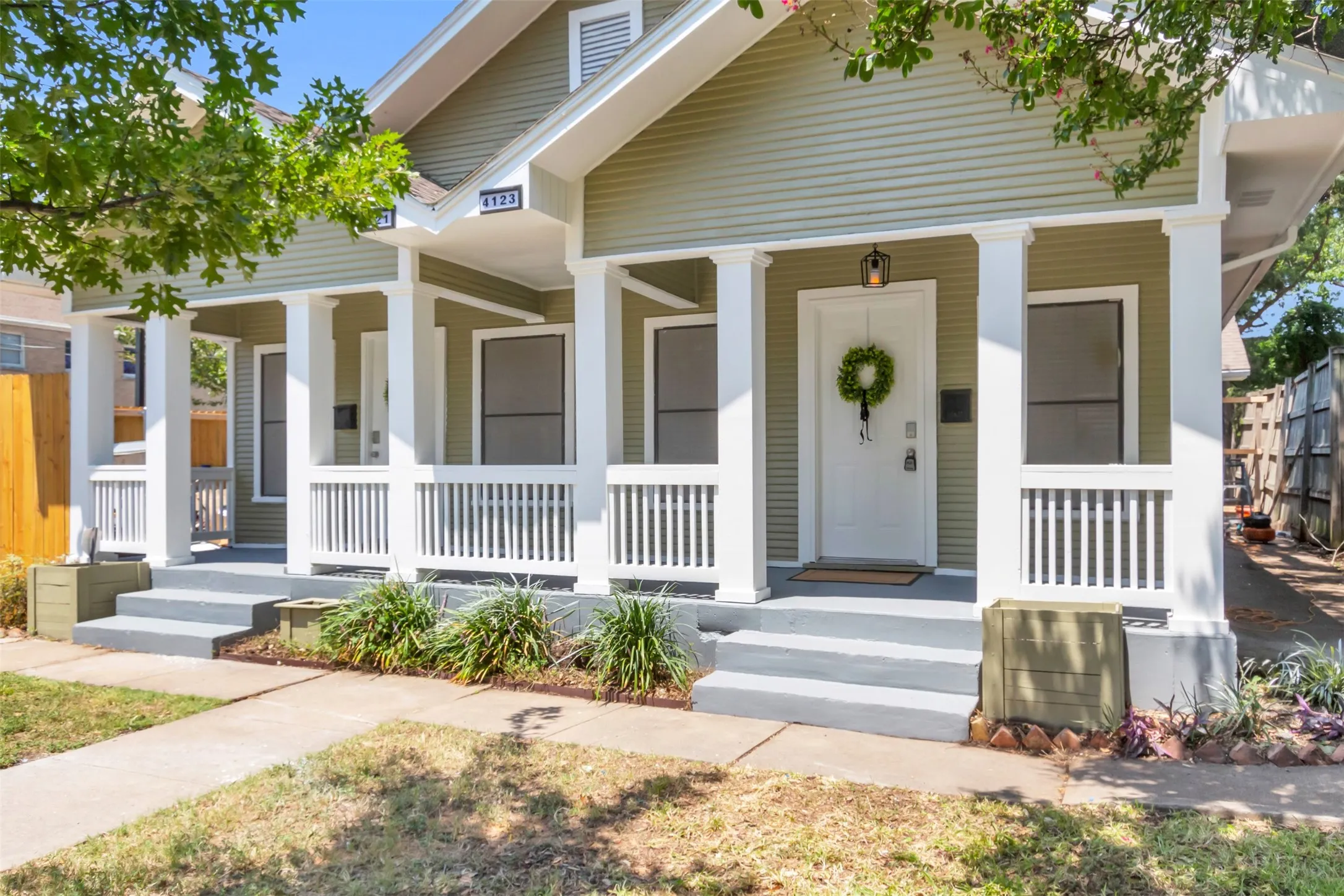 Front exterior featuring light green siding, a covered porch with white columns and railing, a white entry door with an overhead lantern, and gray porch steps