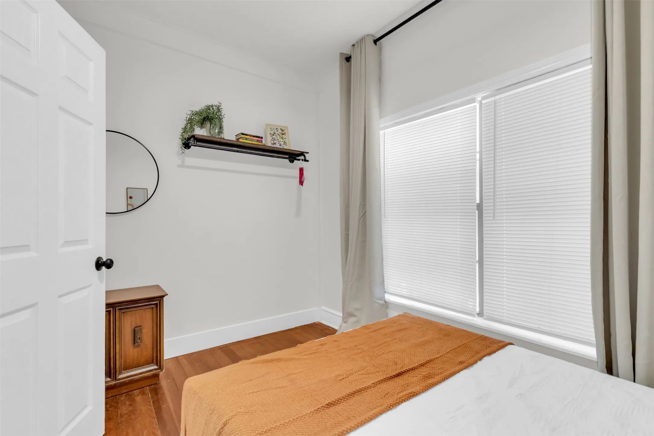 Bright bedroom featuring wood-finish flooring, white paneled door with black hardware, and a large window with privacy blinds