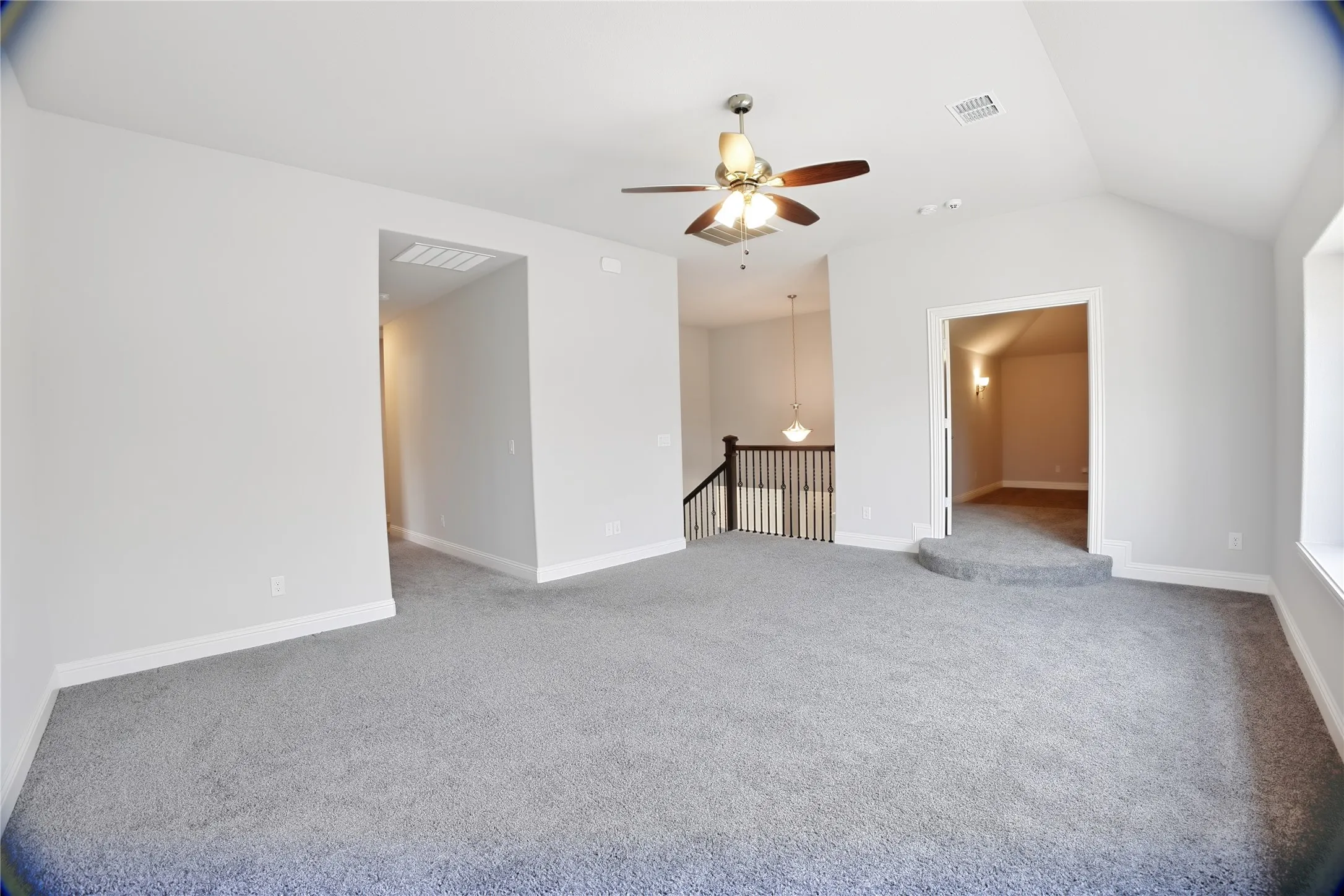 Spacious carpeted room featuring vaulted ceilings, a ceiling fan with integrated lighting, a raised entry to an interior room, and a staircase with wood newel posts and railings