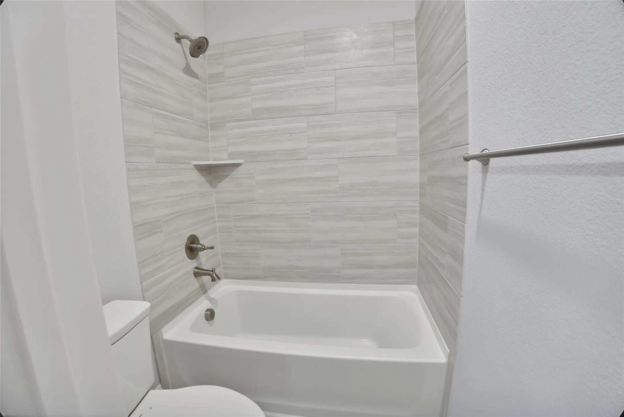 Bathtub and shower combination featuring brushed nickel fixtures, a built-in corner shelf, and light gray horizontal tile surround
