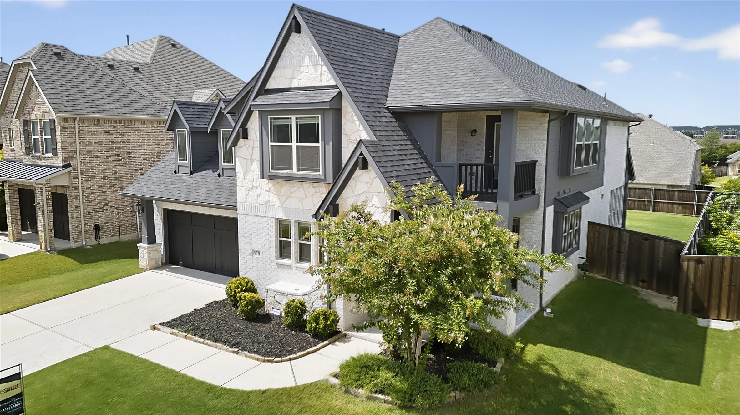 Contemporary two-story exterior featuring white stone and grey siding accents, a dark shingle roof, an attached two-car garage with a dark panel door, and a private second-story balcony