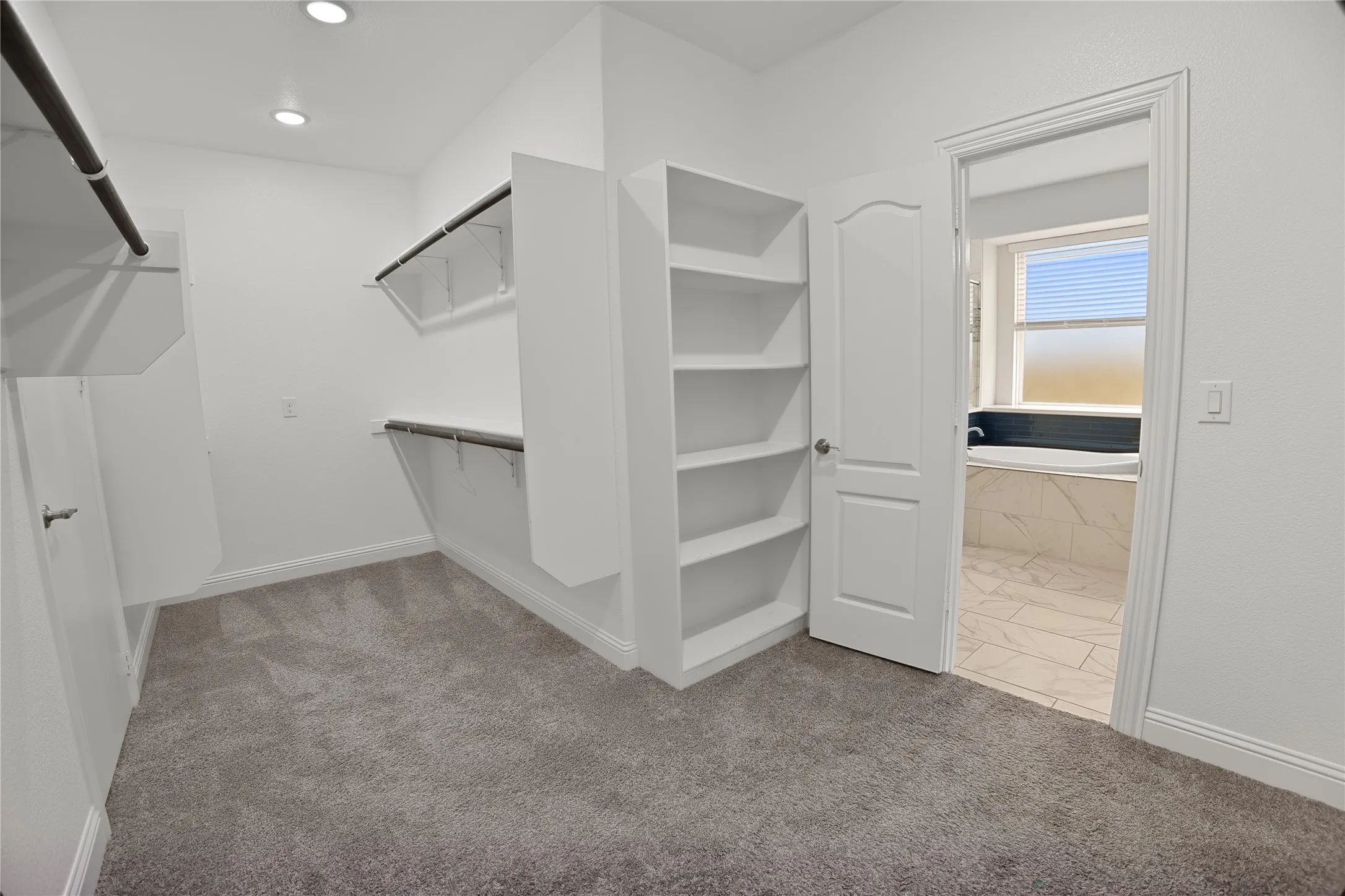 Walk-in closet with carpet flooring, recessed lighting, built-in shelving, and direct access to a bathroom featuring a soaking tub