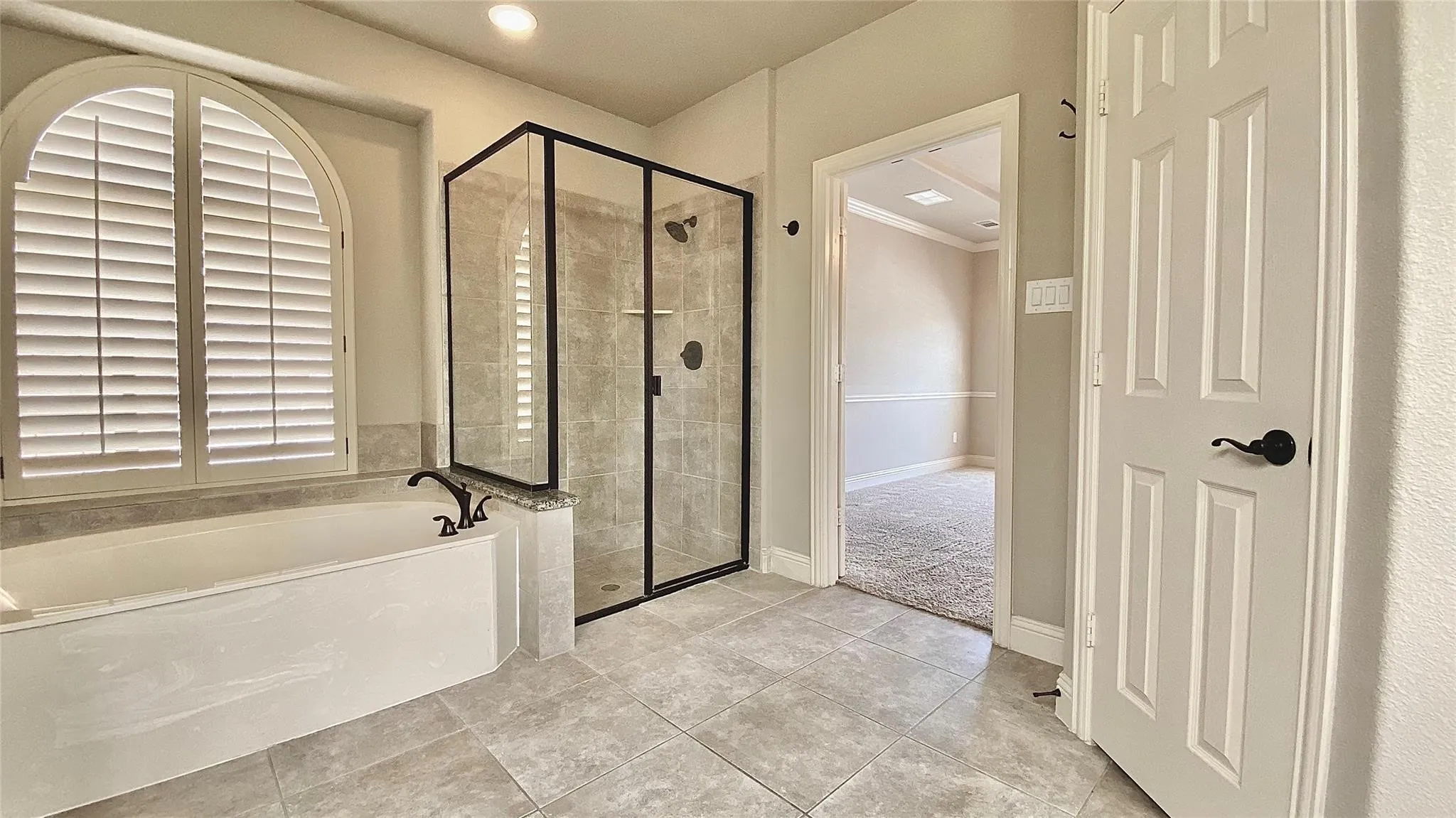 Soaking tub with arch-top plantation shutters, walk-in shower with black-framed glass enclosure, and gray tile flooring
